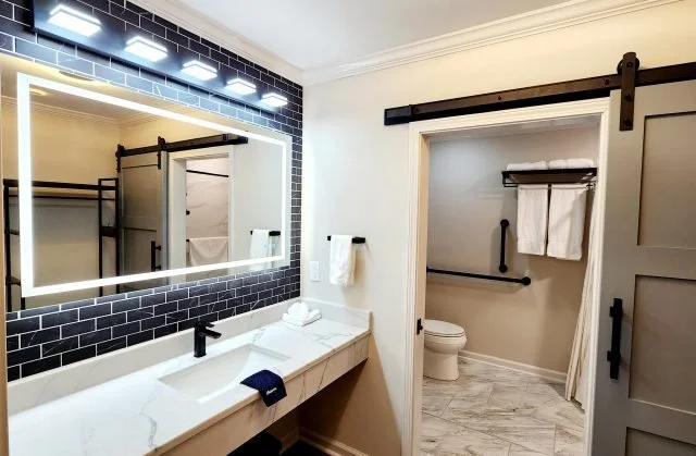 Modern bathroom with a large illuminated mirror above a white marble countertop with a black faucet. To the right, a separate toilet area with a white toilet, towel rack, and a set of white towels, separated by a sliding barn door.