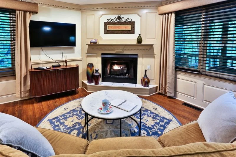 Living room with a fireplace, flat-screen TV, a round coffee table with a mug and notebook, beige curtains, and wooden floors.