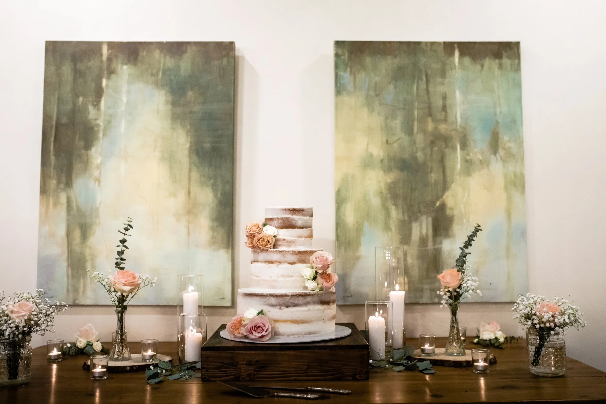 Decorative wedding or event setup with a rustic tiered cake, surrounded by pink and white roses, candles, and greenery, set against two large abstract paintings with green and neutral tones.