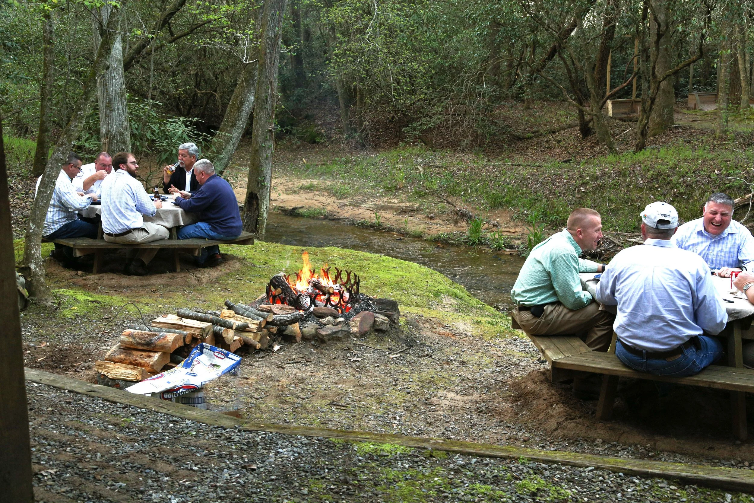 Group of men sitting at picnic tables outdoors in a wooded area, enjoying drinks and laughter near a small campfire over a creek.