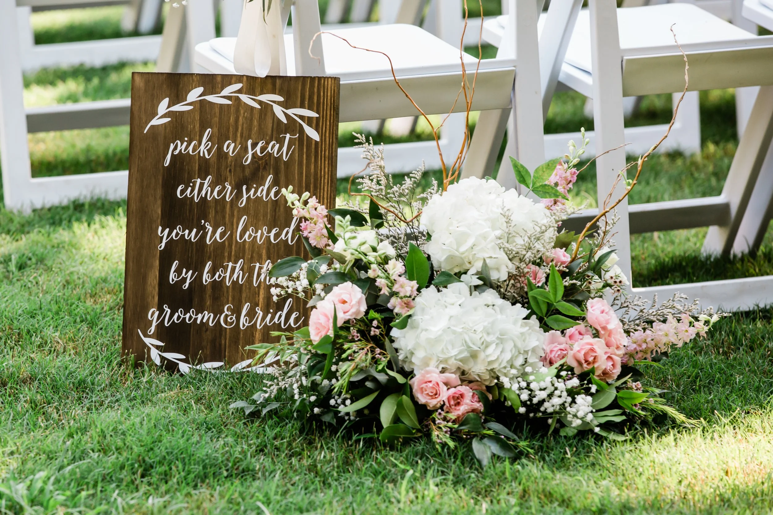 Wedding sign with wooden background and white lettering, surrounded by a floral arrangement of white and pink flowers, set outdoors on green grass, aligned with white chairs in the background.