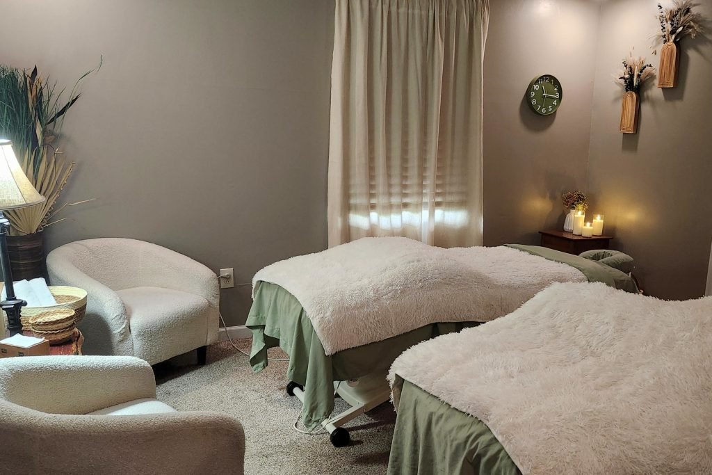 A cozy spa or massage room with two massage beds covered in sheepskin throws, a small table with lit candles, a clock on the wall, a curtain, and a chair.