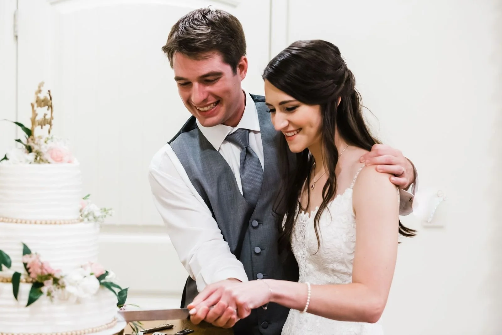 A newlywed couple cutting a wedding cake together, smiling, at their reception.