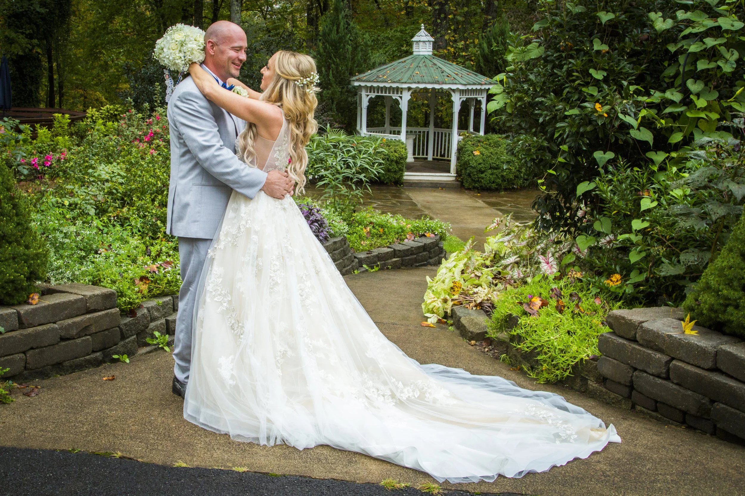Bride and groom dancing outdoors in a garden, surrounded by greenery and colorful flowers, with a decorative gazebo in the background.