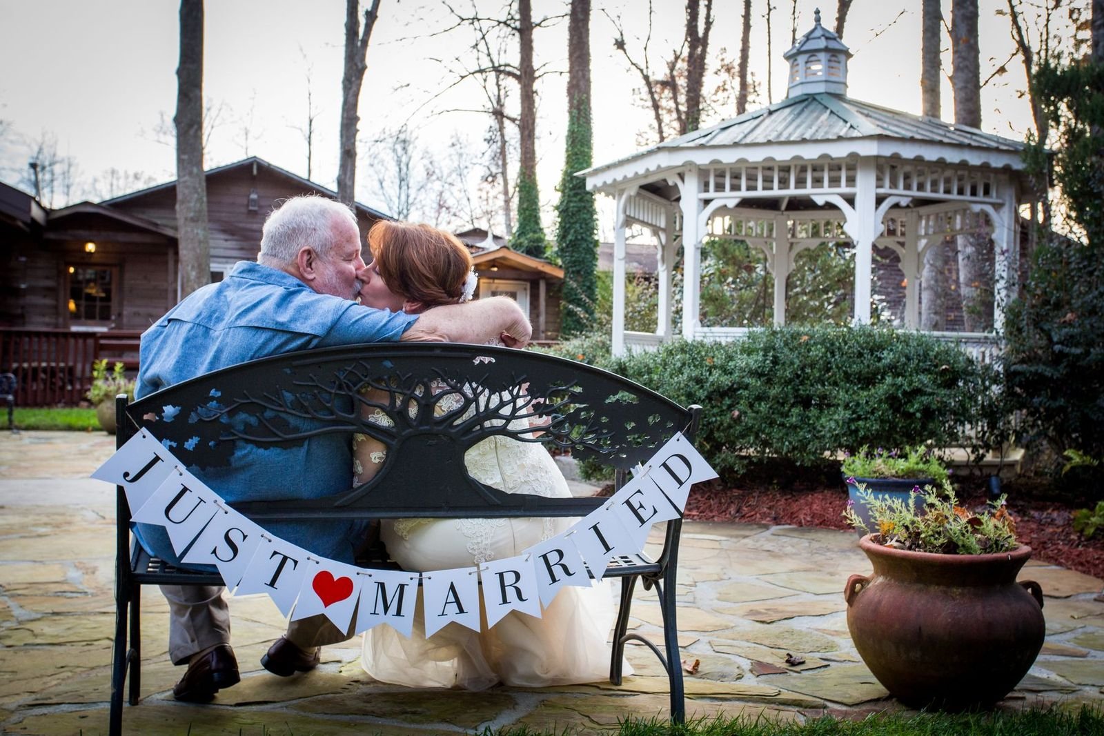 An elderly man and woman sharing a kiss on a decorative outdoor bench with a 'Just Married' banner, in a garden setting with a gazebo and potted plants.