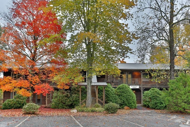 A two-story wooden lodge with a balcony, surrounded by colorful autumn trees with red, orange, and yellow leaves, and some green foliage, in a parking lot