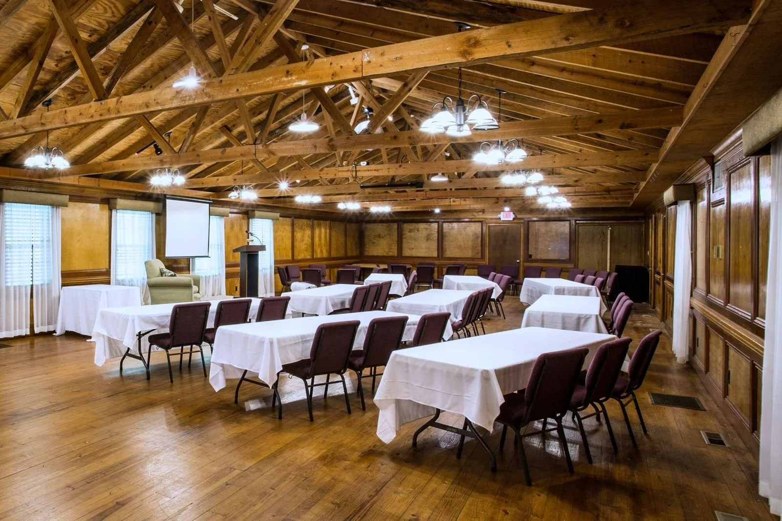 Empty conference room with arranged tables covered with white tablecloths, purple chairs, wooden walls, a projector screen, a podium, and ceiling lights.