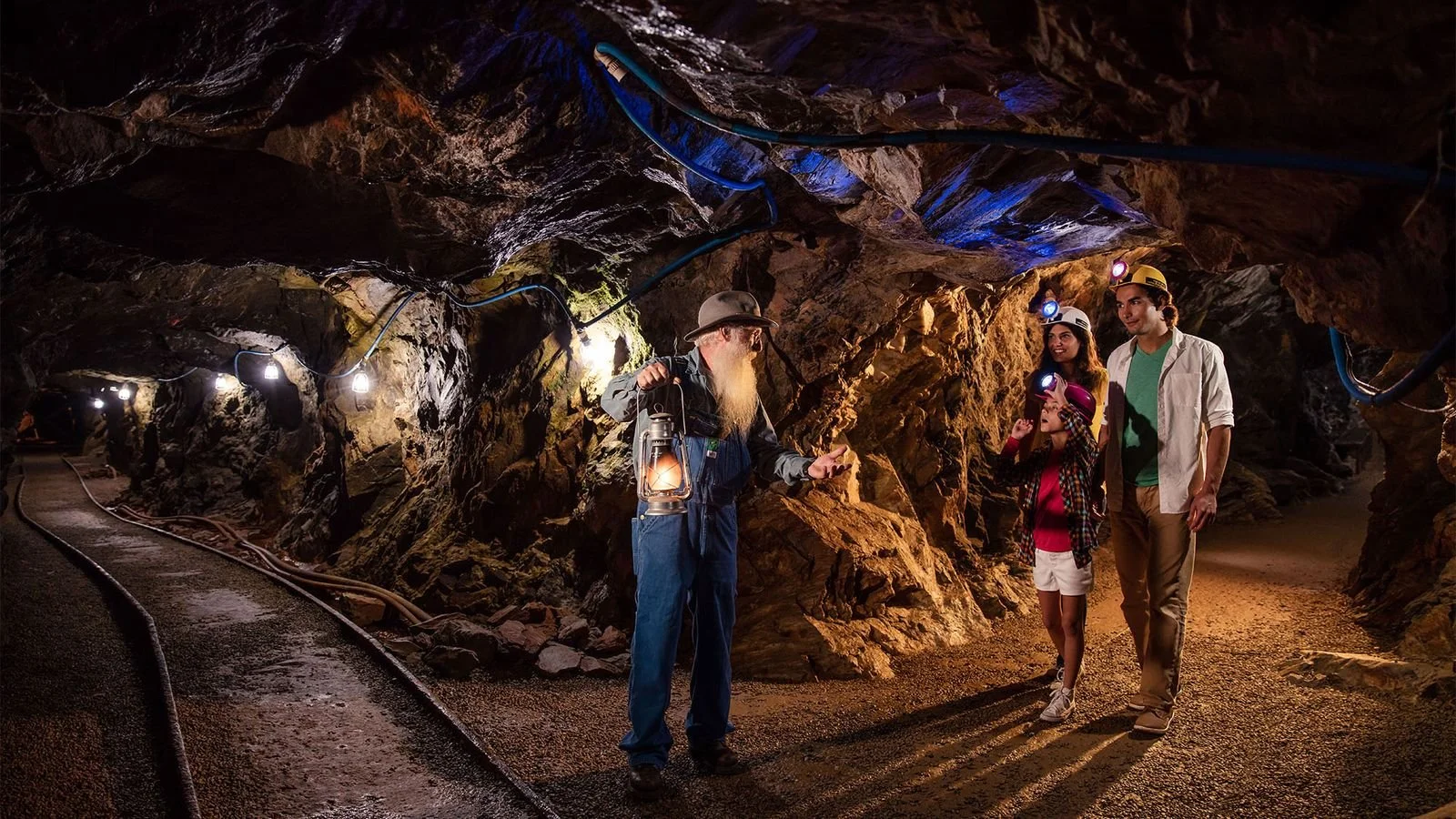 A group of three young explorers, wearing helmets with headlamps, walk through a mining tunnel with a miner, who is wearing a helmet and holding a lantern, in the cave. The tunnel is illuminated by electric lights and has rugged, rocky walls.