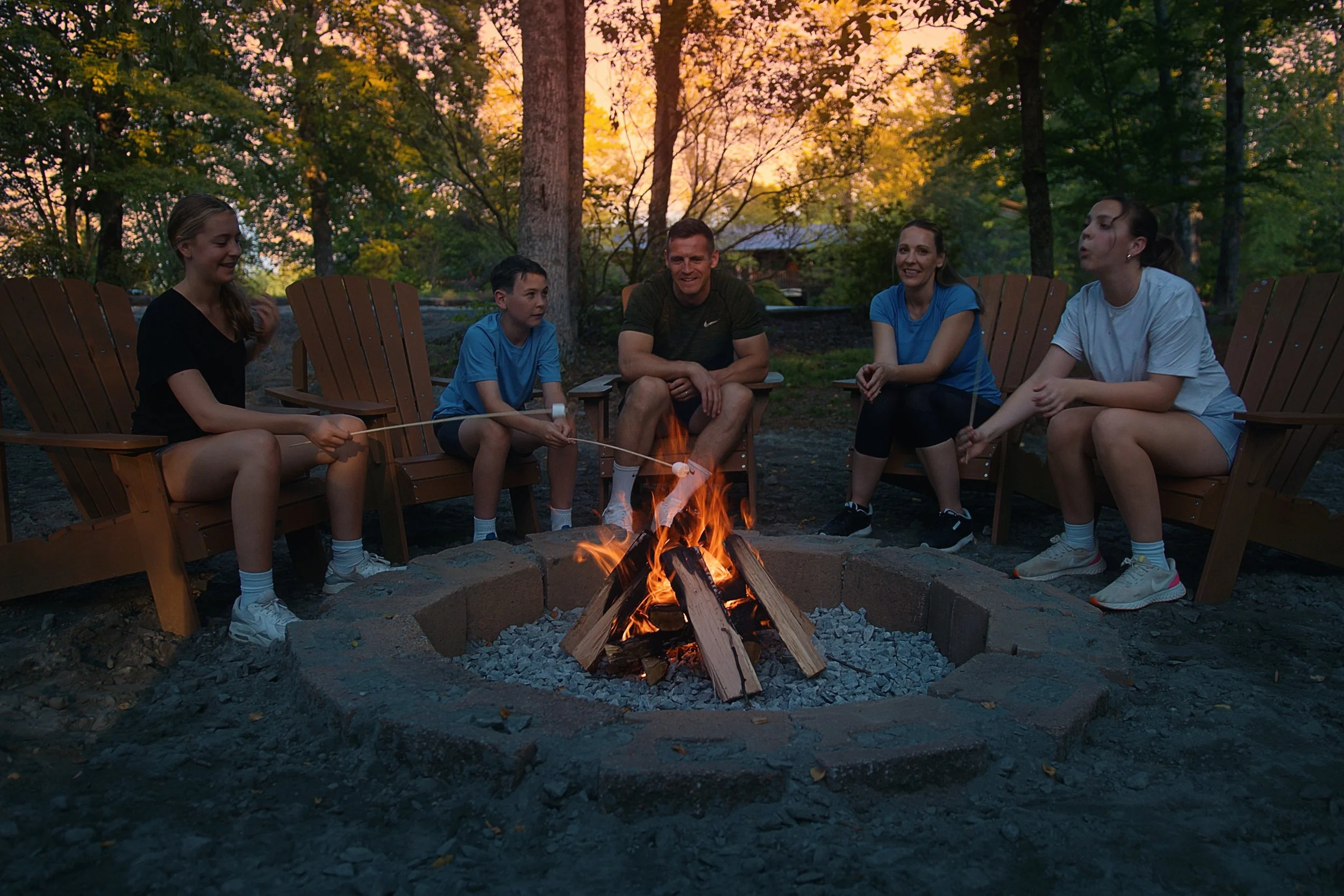 Group of five people sitting on Adirondack chairs around a campfire in a wooded outdoor setting during sunset, engaging in conversation and roasting marshmallows.