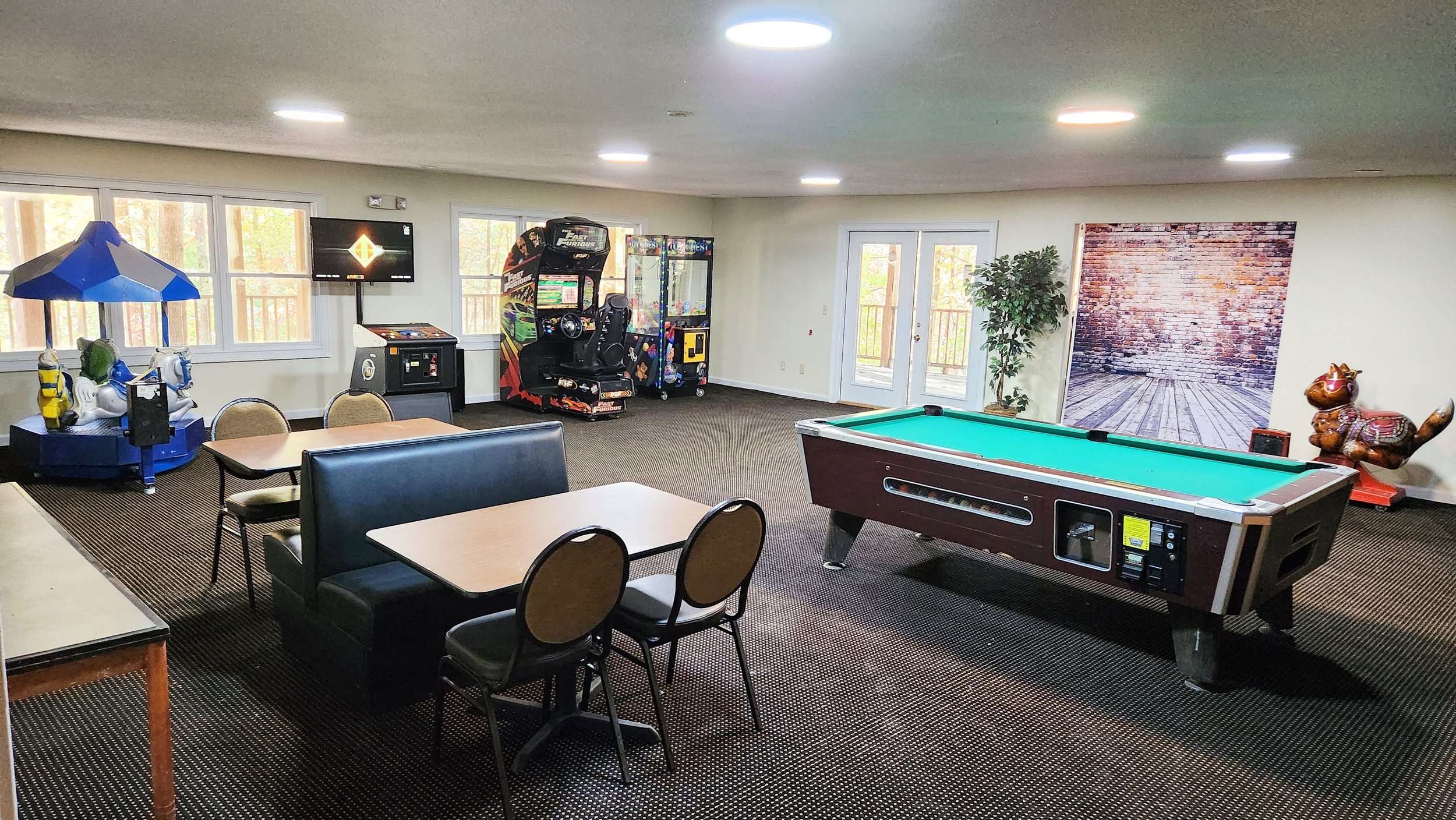 An indoor recreational room with arcade games, a pool table, and children's rides, featuring windows, a wall with a brick backdrop, and a door leading outside.