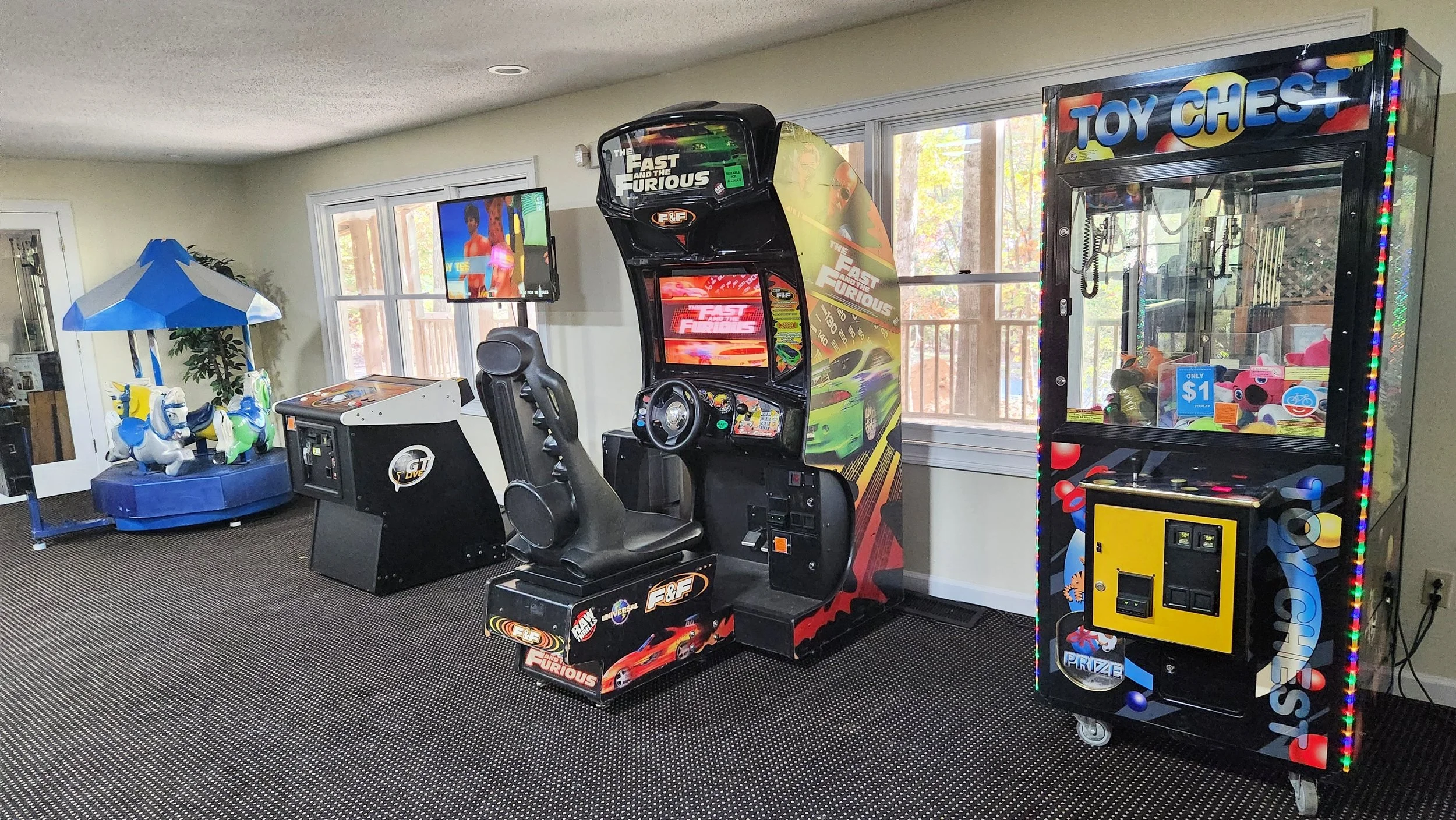 Indoor arcade room with a racing game, claw machine, coin-operated horse carousel, and a flat-screen TV.