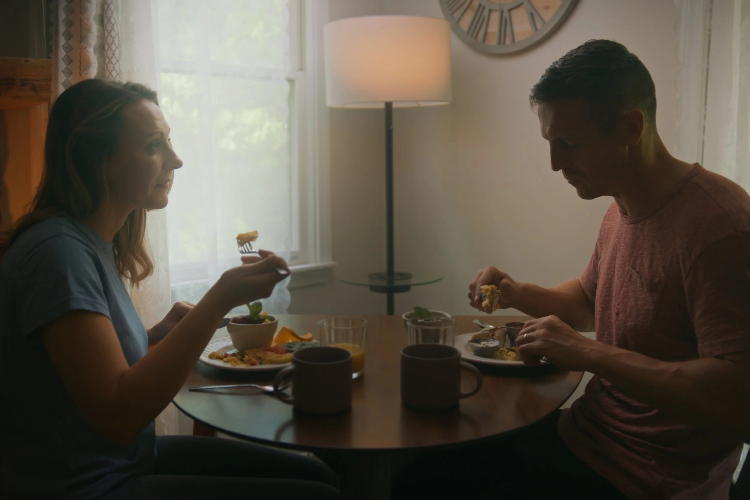 A woman and a man eating dinner together at a table, with a window and a large clock on the wall behind them. The woman has long hair and is holding a fork, while the man has short hair and is looking at his plate.