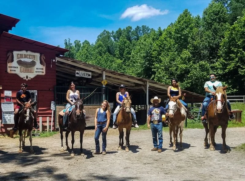 Group of six people, some on horseback, at a ranch with trees and a blue sky in the background.
