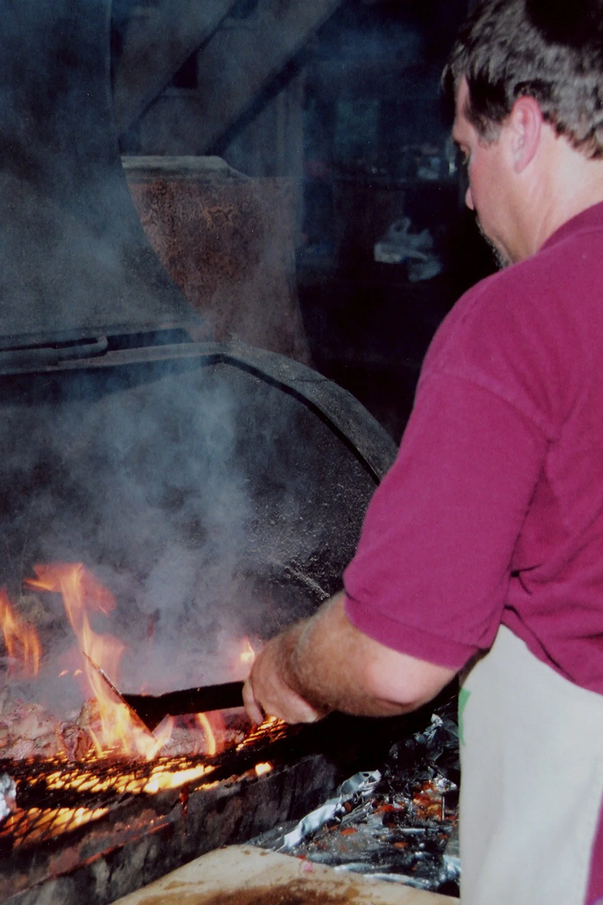 A man in a burgundy shirt and white apron tends a large outdoor charcoal grill that is on fire, with flames and smoke rising from the grill.