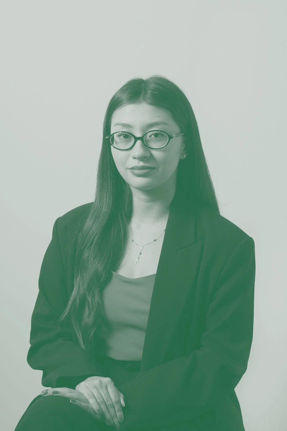 Retrato de uma mulher usando óculos, blusa cinza, blazer preto, acessório de corrente delicado, sentada na posição de três quartos contra fundo branco.