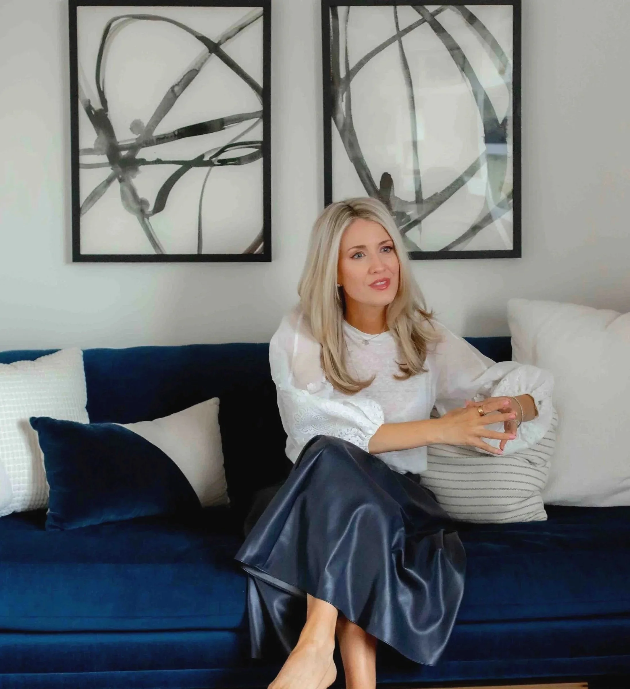 A woman sitting on a sofa with a blue cushion, wearing a white blouse and a black leather skirt, with her hands clasped on her lap. Behind her are two black-framed abstract black and white paintings hanging on a white wall. A black purse and a navy blue jacket are placed on the sofa.