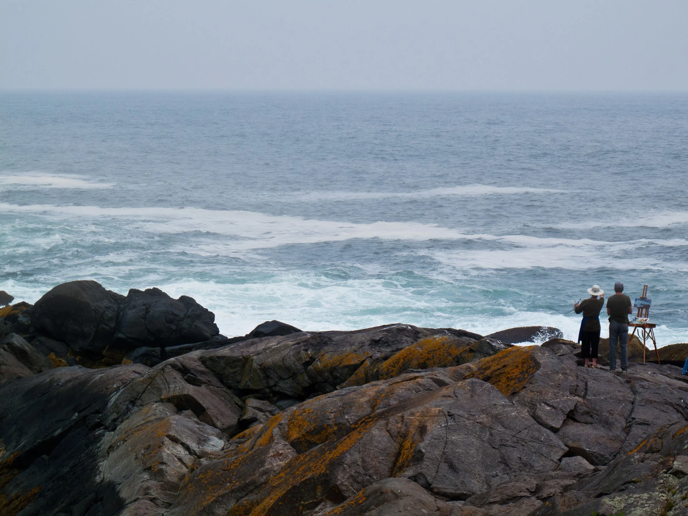 Two people standing on rocks by the ocean, one taking a picture or fishing, in front of the water with waves and a hazy sky.