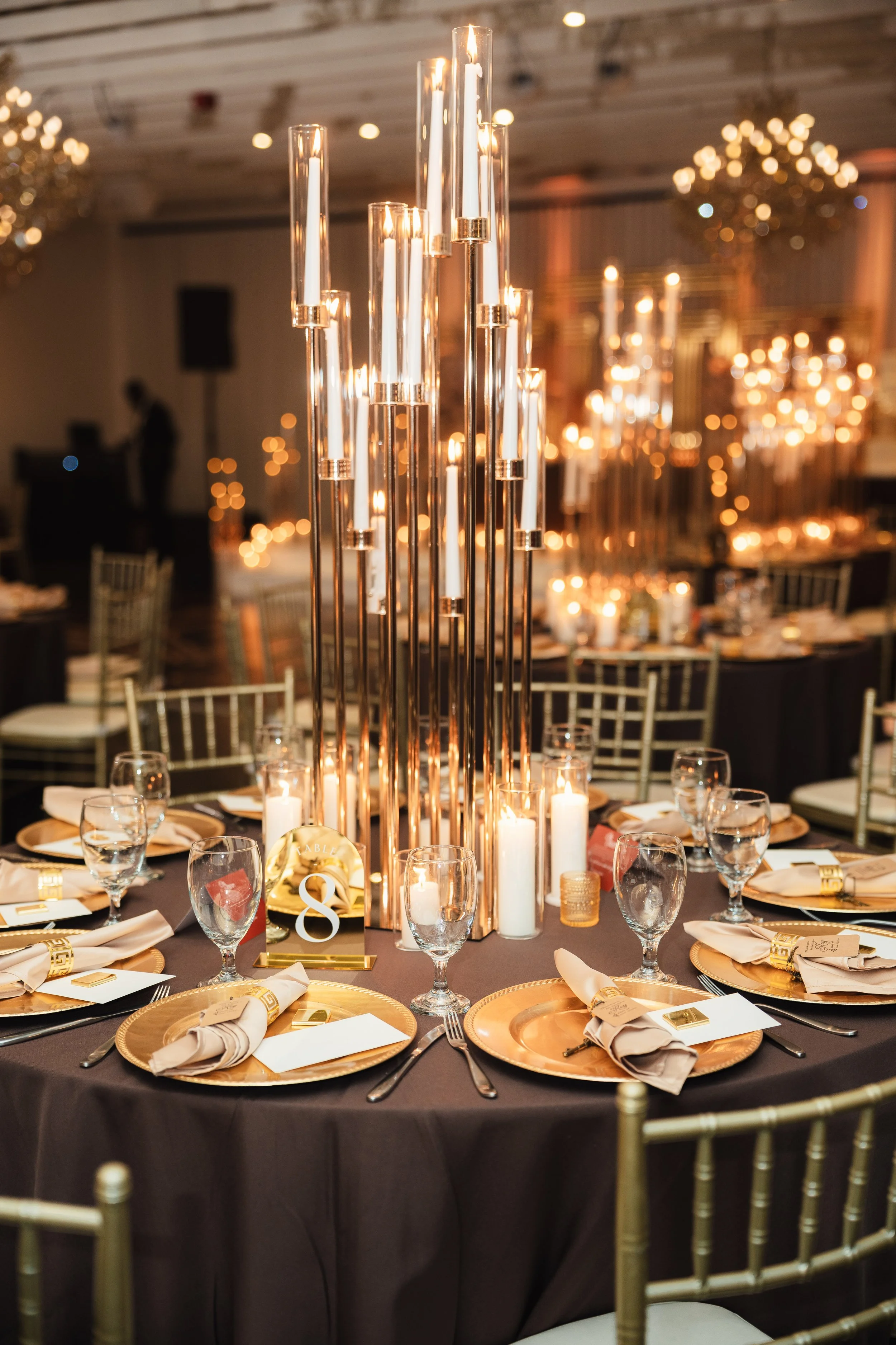 Elegant banquet table with gold chargers, beige napkins, glassware, and a tall centerpiece with cylindrical candle holders and lit candles, in a decorated event hall.