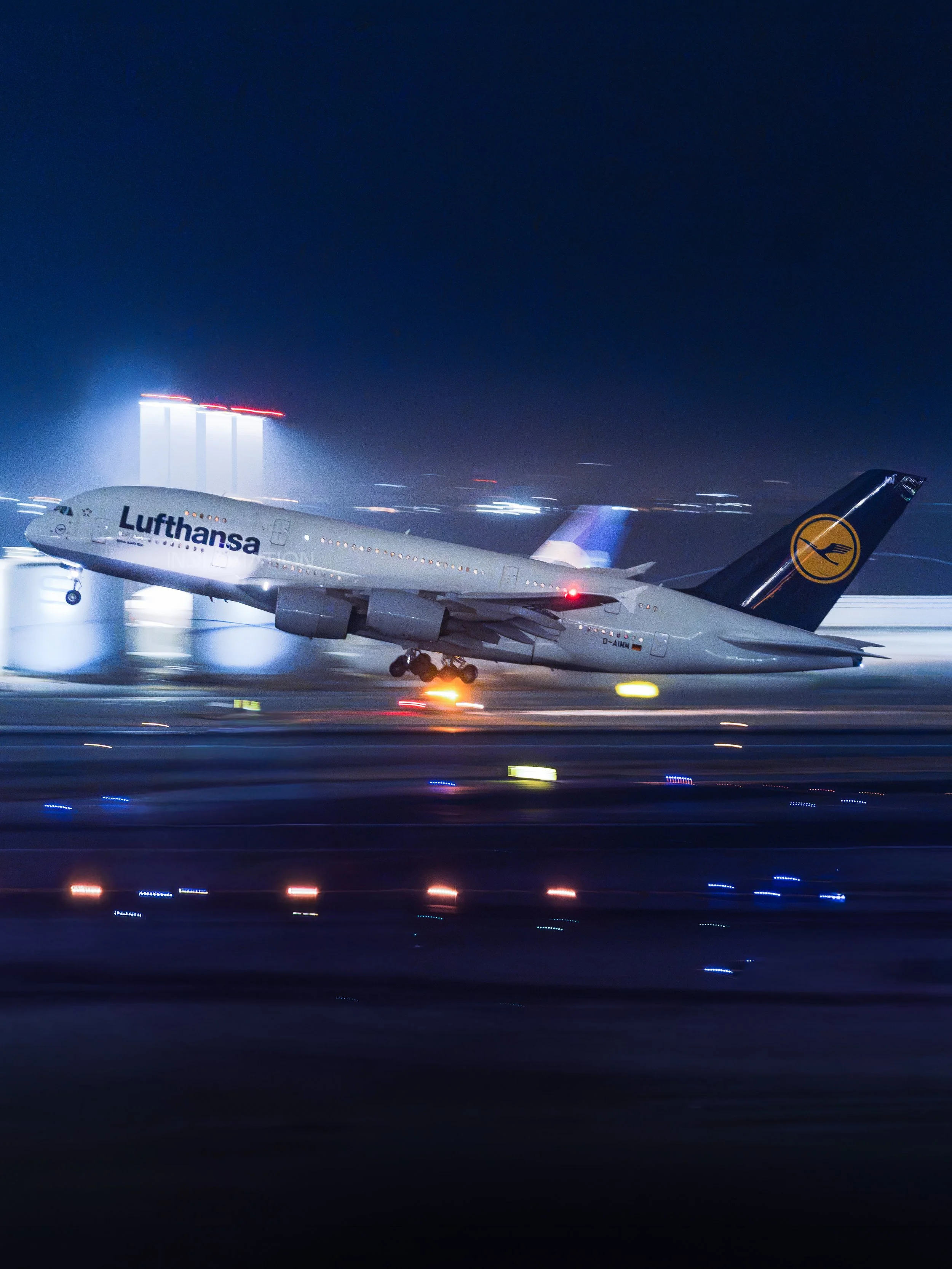 A Lufthansa commercial airplane on a runway at night, with the city skyline and tall buildings in the background. The scene is illuminated by lights, creating a motion blur effect.