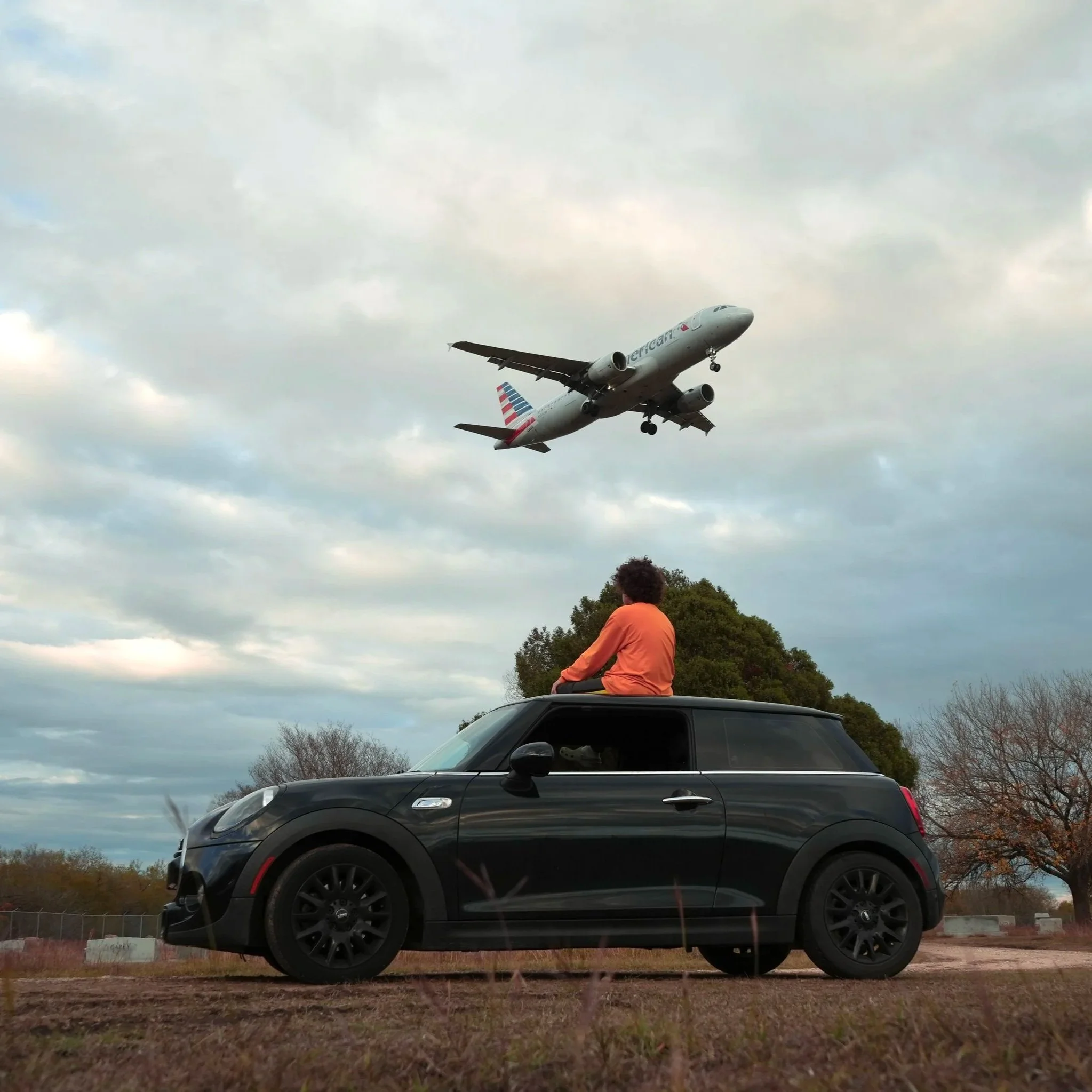 A black SUV parked on dirt, with a person in an orange shirt sitting on top of the vehicle, while an airplane is flying low in the cloudy sky above.