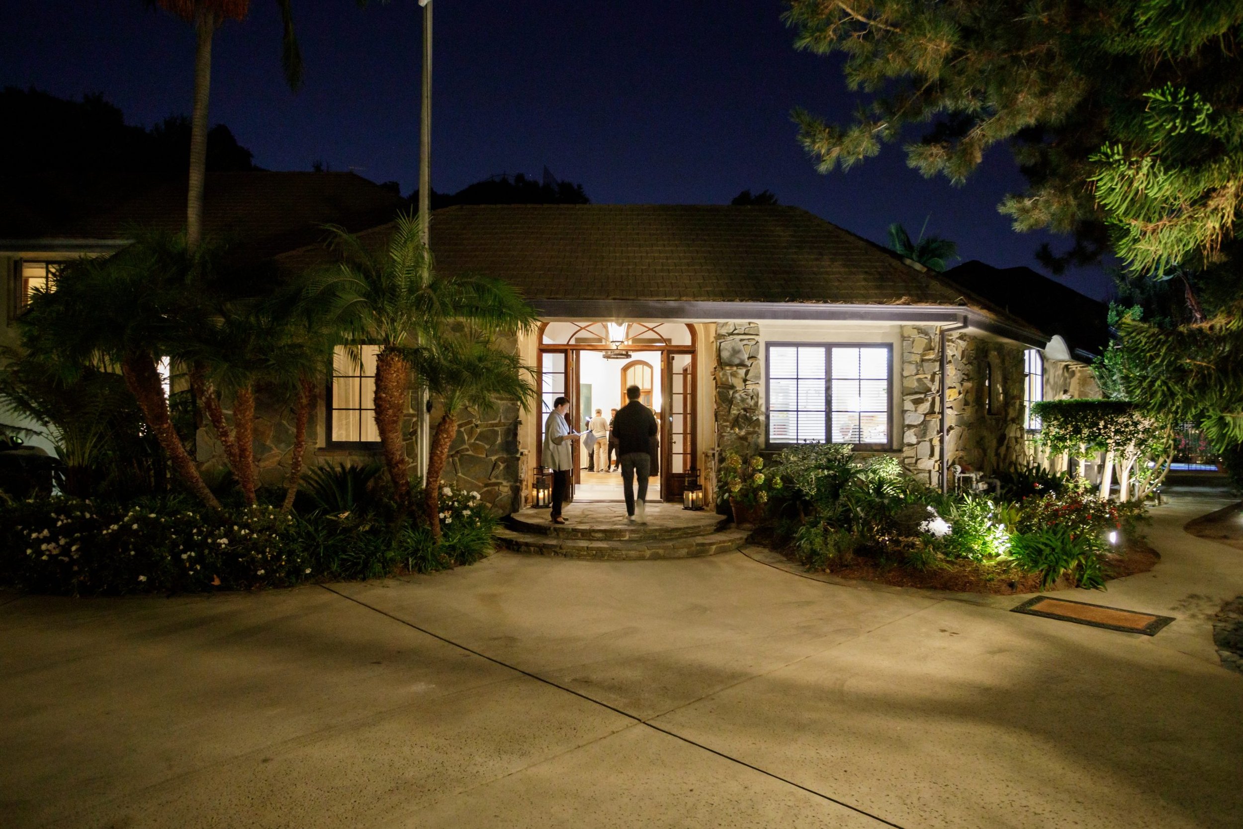 Night view of a house with a stone facade, illuminated open front doors, and large windows. There are palm trees and lush plants in the front yard. People are entering the house, and the surrounding area is lit with outdoor lights.