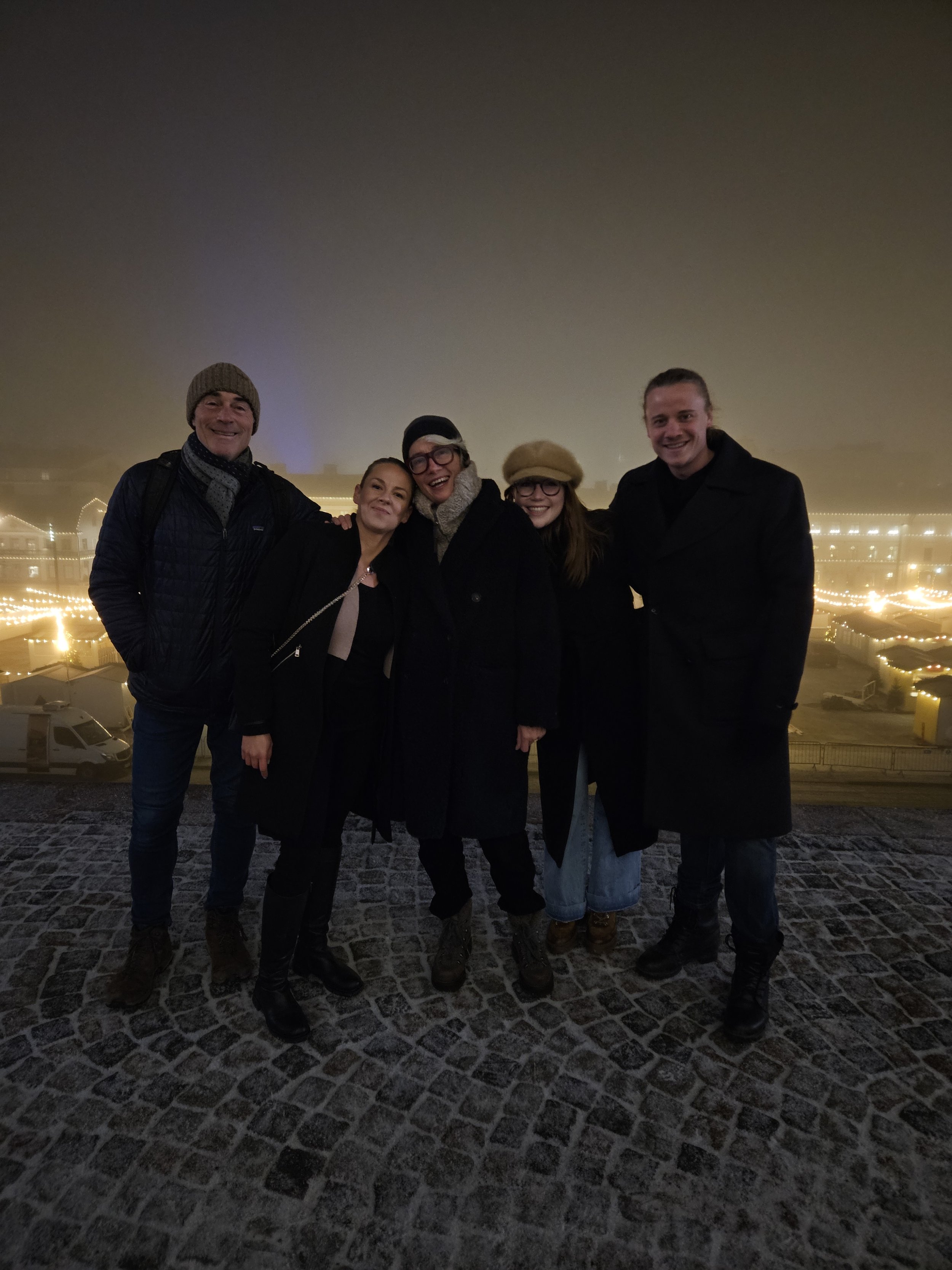 Five people stand together outdoors at night with Christmas lights in the background. They are dressed warmly and smiling.