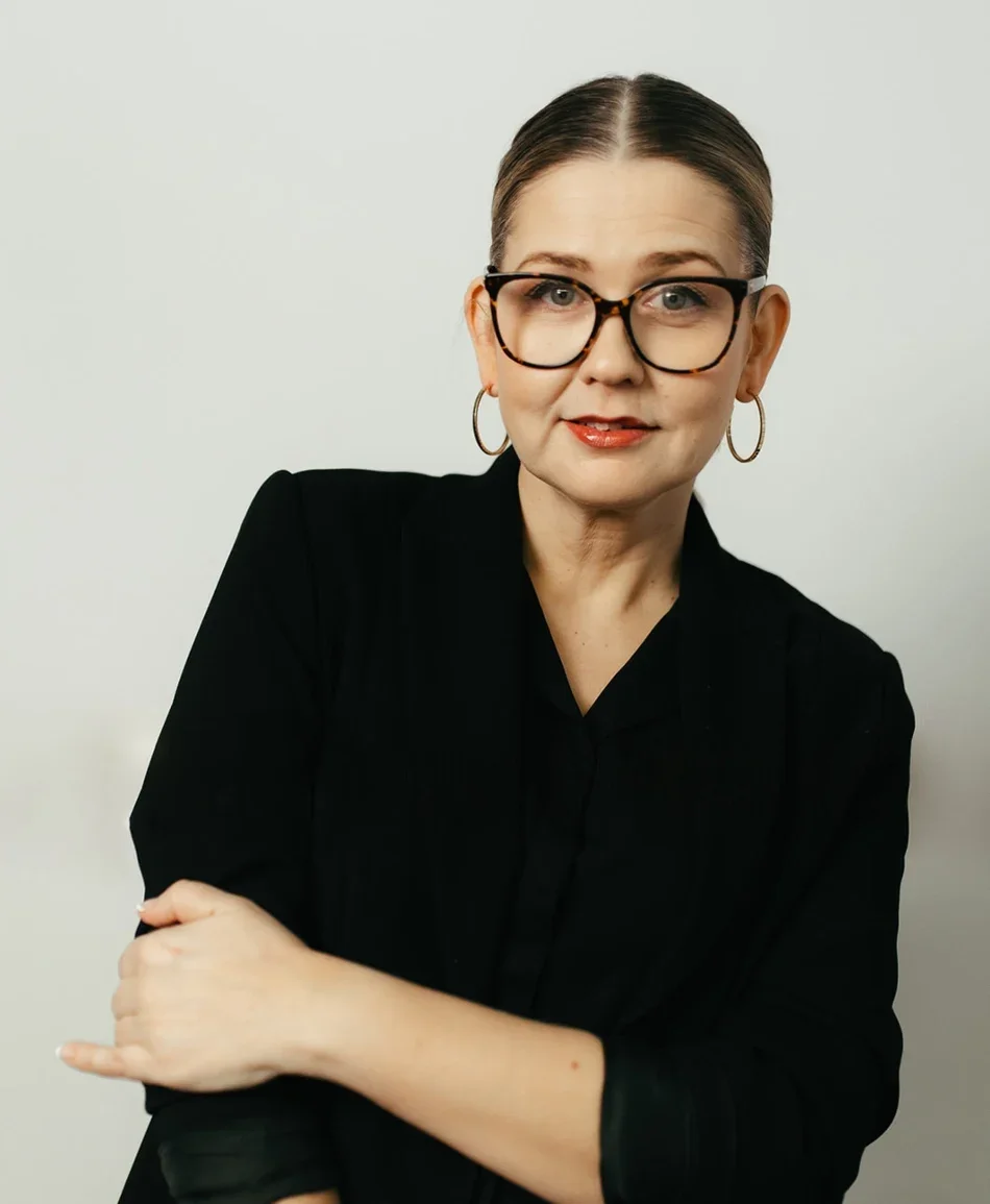 A woman with glasses and hoop earrings, wearing a black blazer, posing with her arms crossed against a plain background.