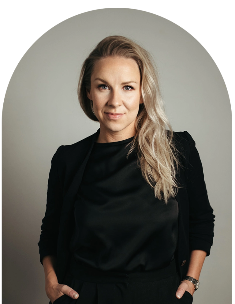 A woman with blonde hair and light skin, wearing a black blazer and blouse, standing against a plain light gray background.