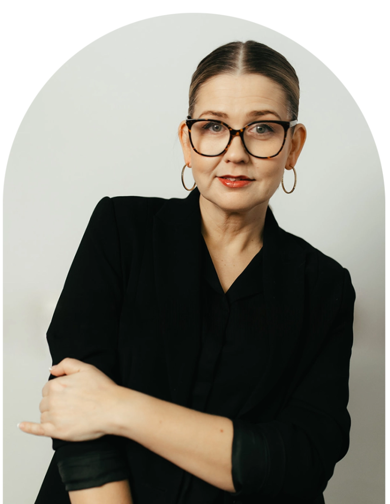 A woman with glasses and hoop earrings, wearing a black blazer, standing against a plain white background.