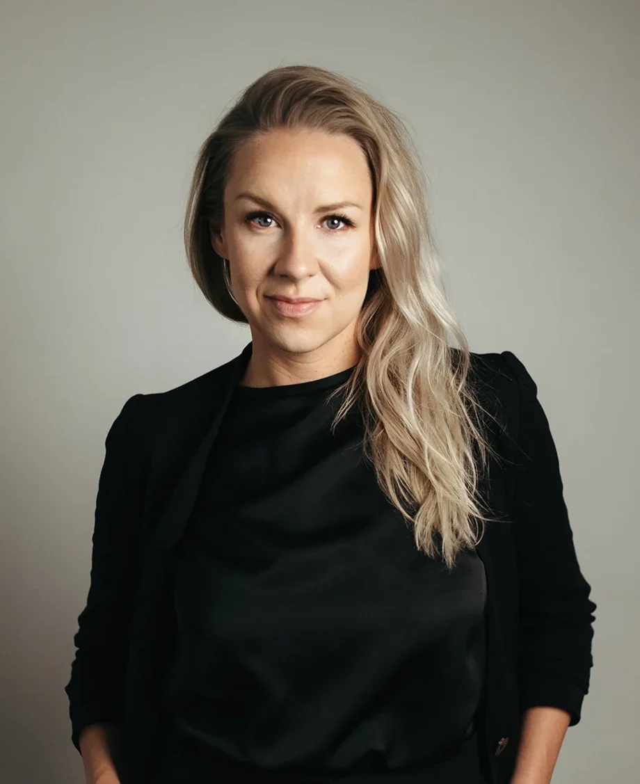 A woman with long blonde hair wearing a black top and blazer, standing against a plain gray background.