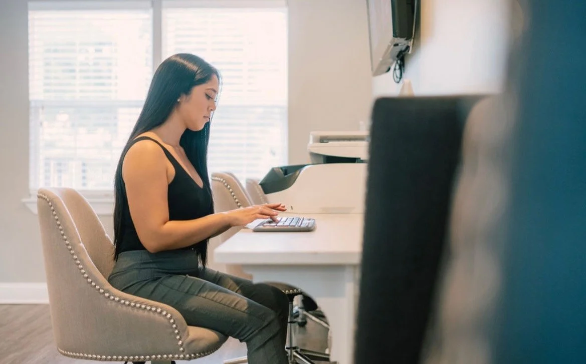A woman with long dark hair sitting at a white desk, using a calculator, in a room with large windows and blinds.