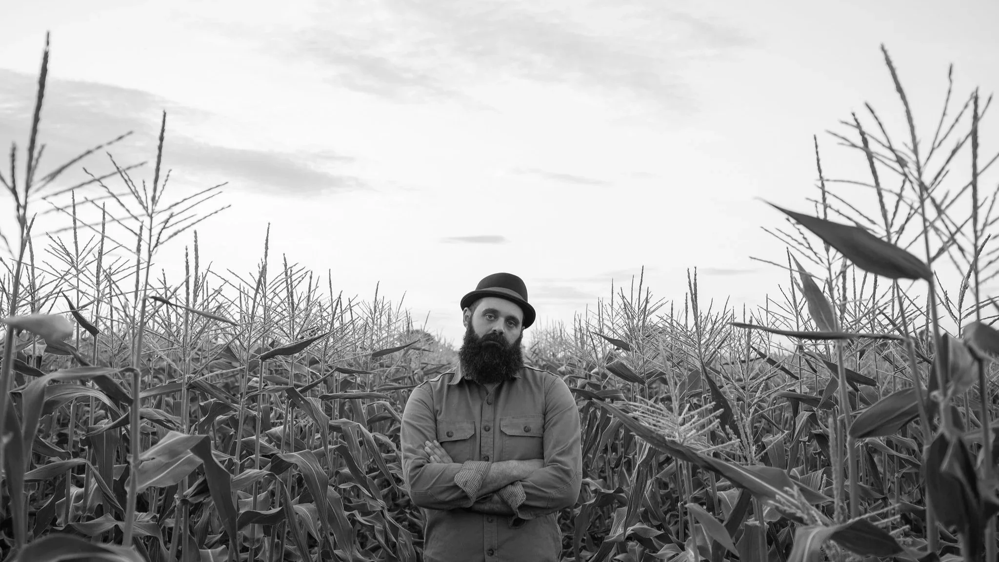 Bernard John wearing a porkpie hat and a button-up shirt standing in a tall cornfield.