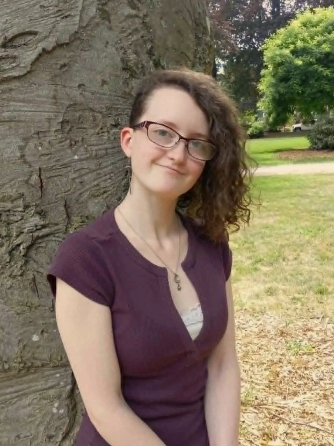 A young woman with curly brown hair and glasses standing outdoors next to a large tree, with green grass and trees in the background.