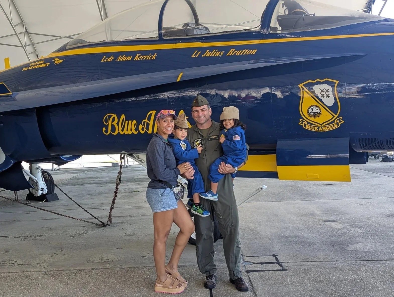A family of four, including two children in blue flight suits, pose for a photo in front of a blue and yellow fighter jet in an aircraft hangar. The family members are smiling, with the mother and father holding the children.