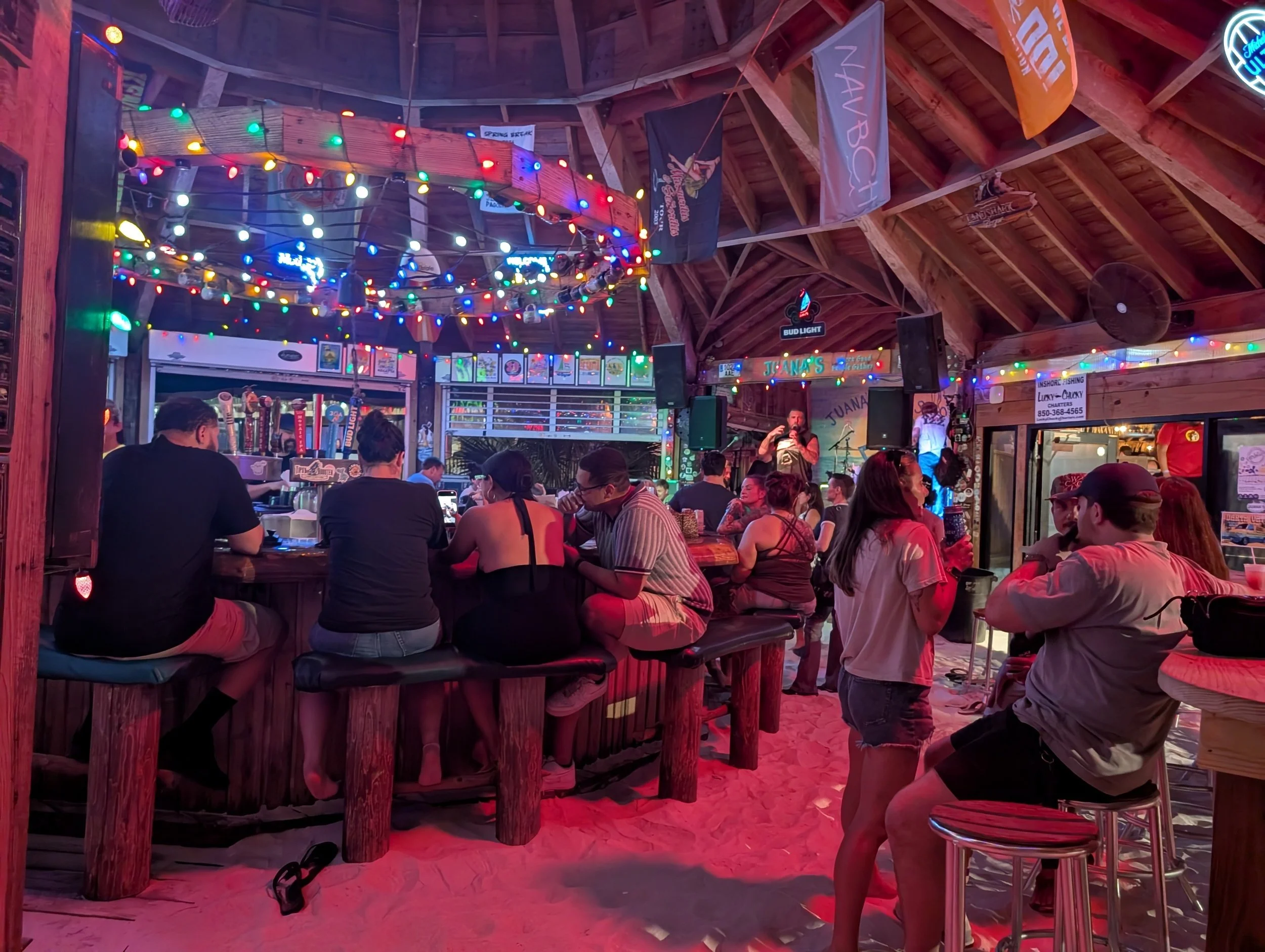 A lively bar and restaurant with colorful string lights hanging from the wooden ceiling, filled with people sitting at tables and standing, enjoying drinks and conversation. A man is speaking into a microphone, suggesting live entertainment, with a bartender visible behind the bar. Neon signs and flags are displayed on the walls