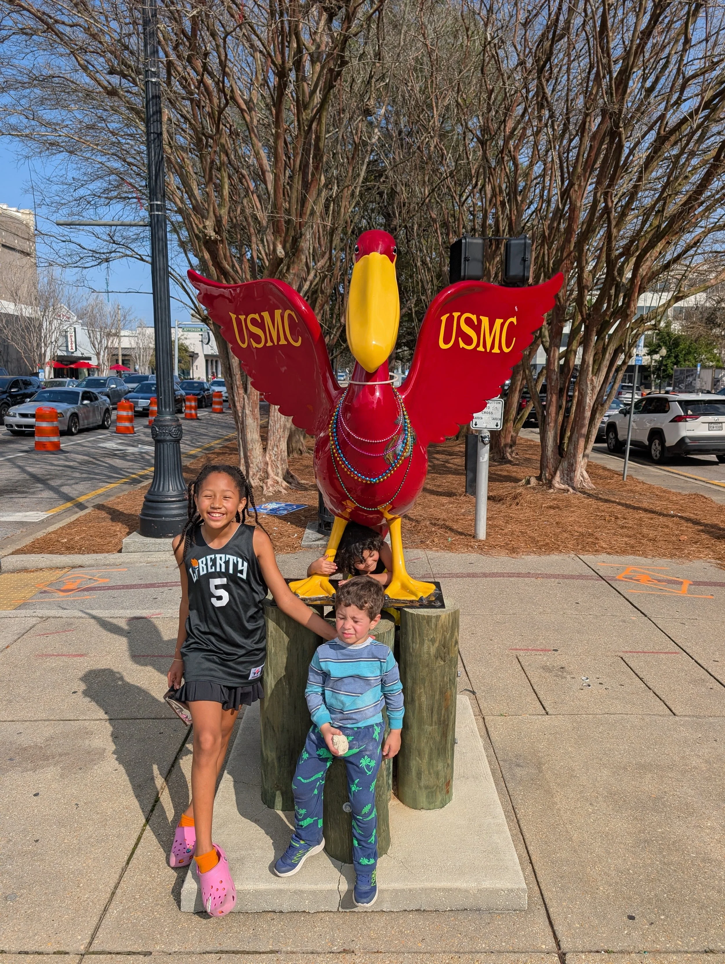 Children standing in front of a red USMC mascot statue decorated with beads, outdoors on a sidewalk with trees and parked cars in the background.