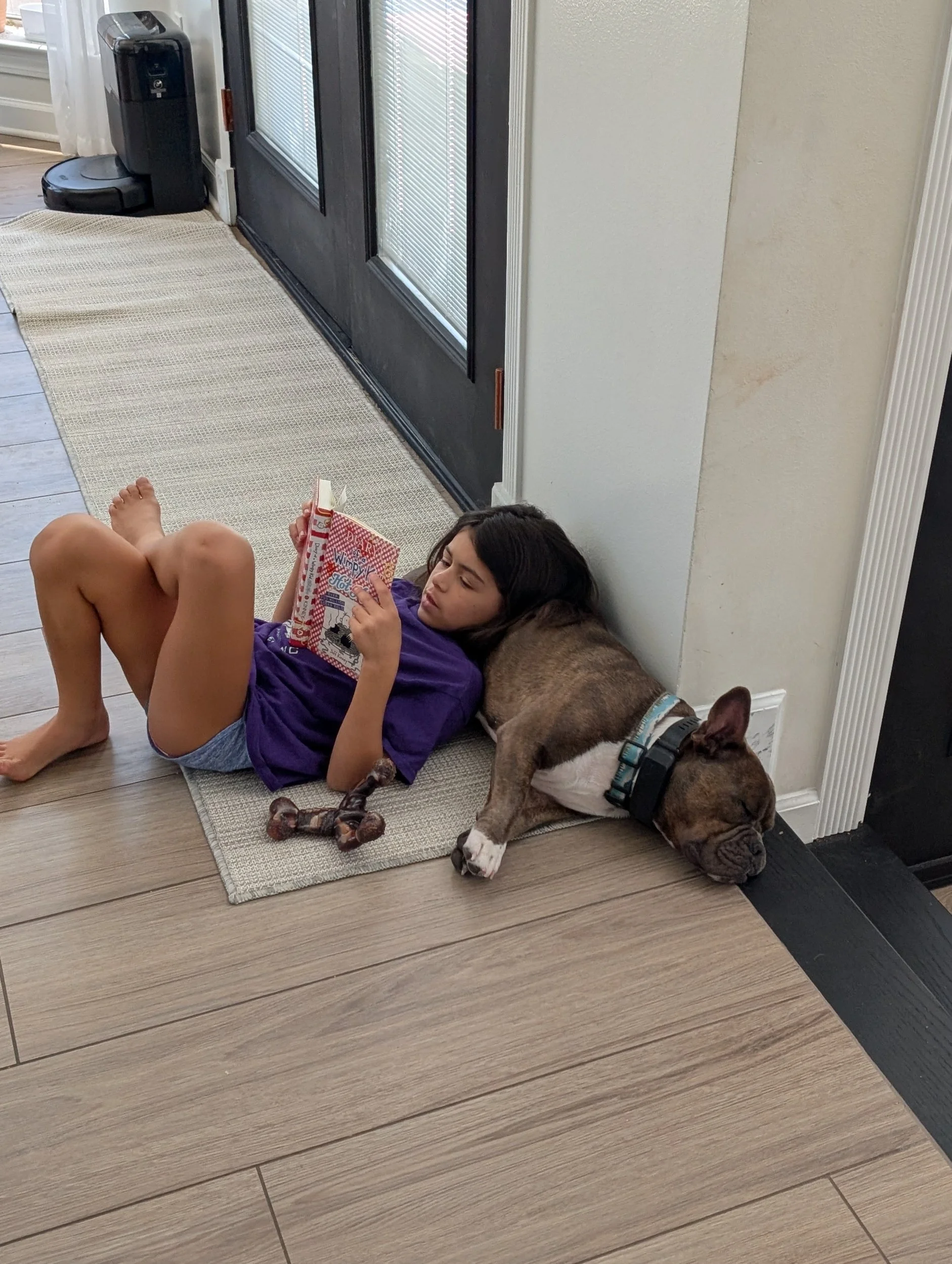 A girl lying on the floor reading a colorful book with her dog resting beside her on the wooden floor near a wall and door in a home interior.