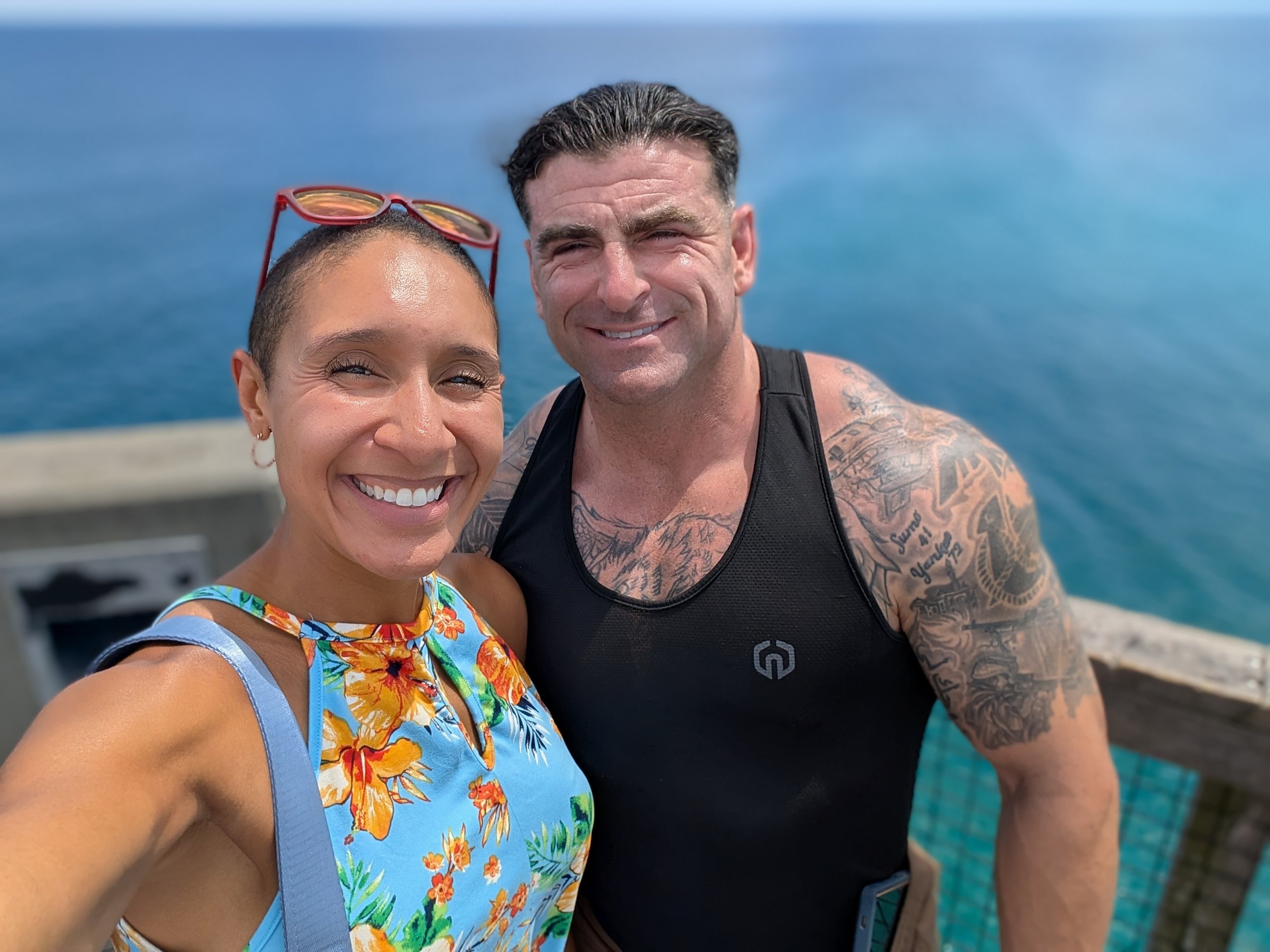 A woman and a man taking a selfie together outdoors near the ocean, smiling at the camera. The woman has short hair, sunglasses on her head, and is wearing a colorful floral top. The man has short dark hair, tattoos on his arms and chest, and is wearing a black tank top.
