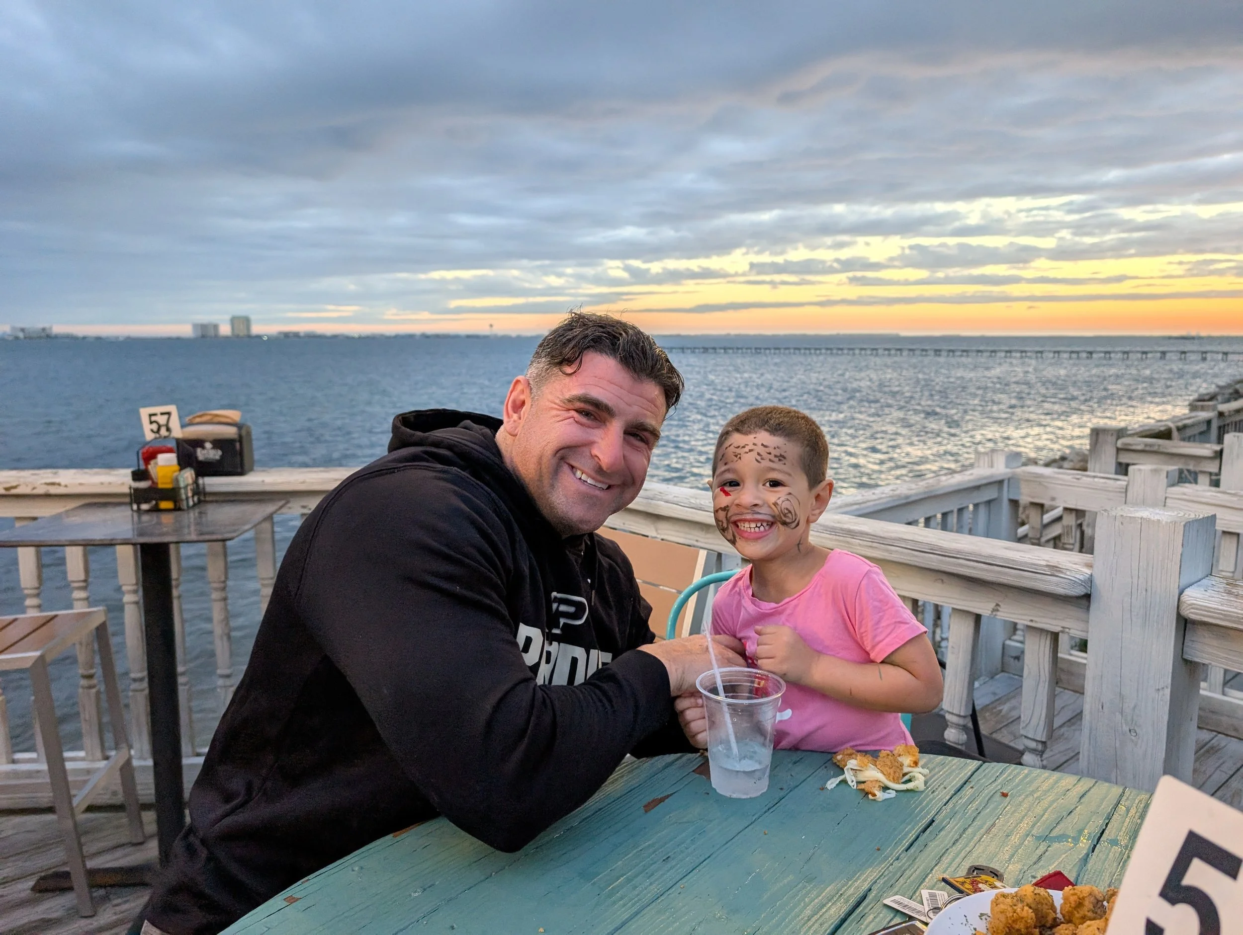 A father and son with face paint smiling at the camera while sitting at an outdoor wooden table near water during sunset in Navarre Florida. The boy is wearing a pink shirt, and the man is wearing a black hoodie.