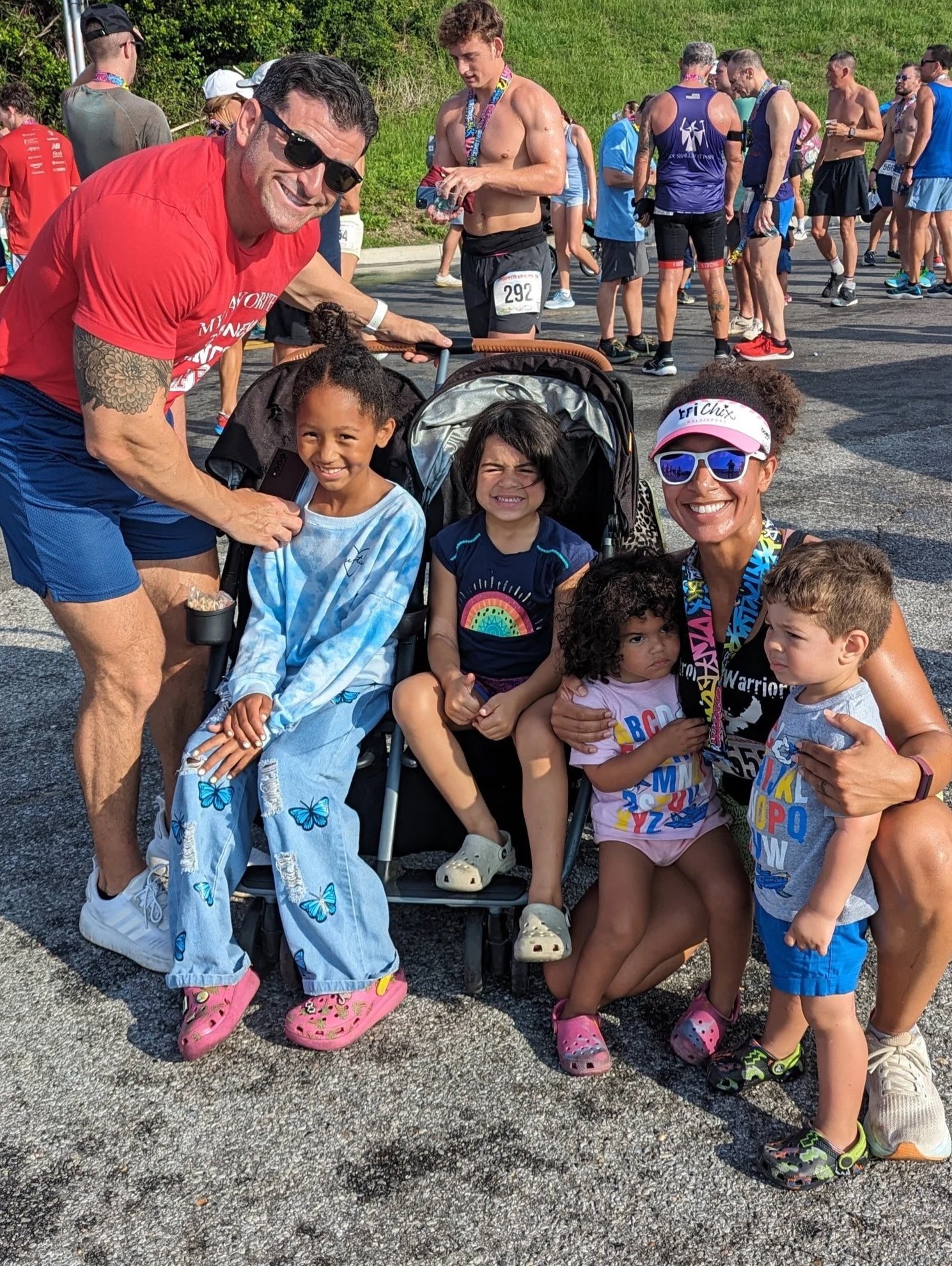 Group of people gathered outdoors on a sunny day, some wearing athletic apparel, at a race event. Three young girls are sitting with two adults, all smiling and enjoying the moment. The background shows more participants and a grassy hill.