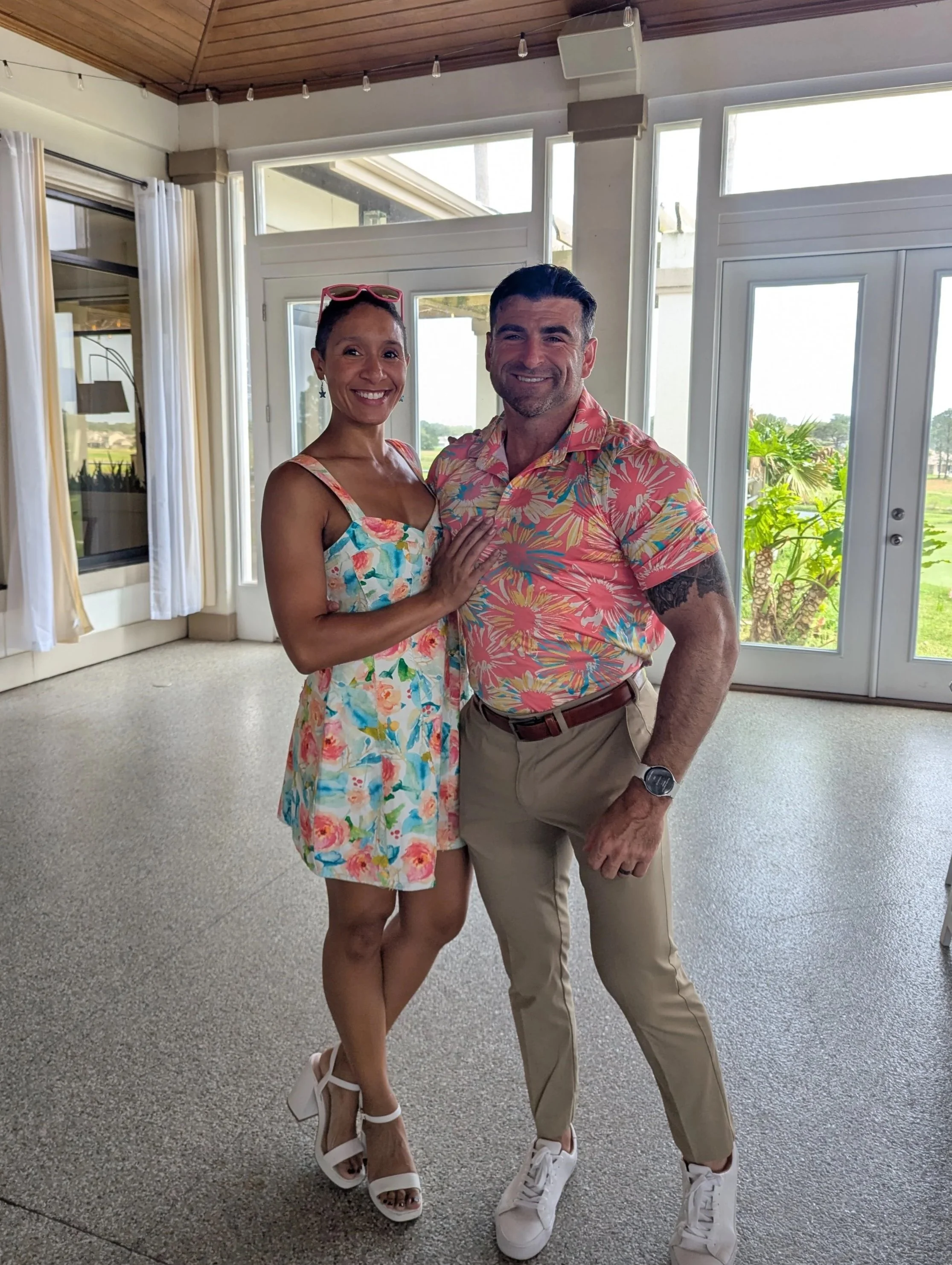 A smiling woman wearing a floral dress and white heels standing next to a smiling man in a pink tropical shirt, khaki pants, and white sneakers inside a bright room with large windows and white curtains at The Pointe, in Gulf Breeze, Florida.
