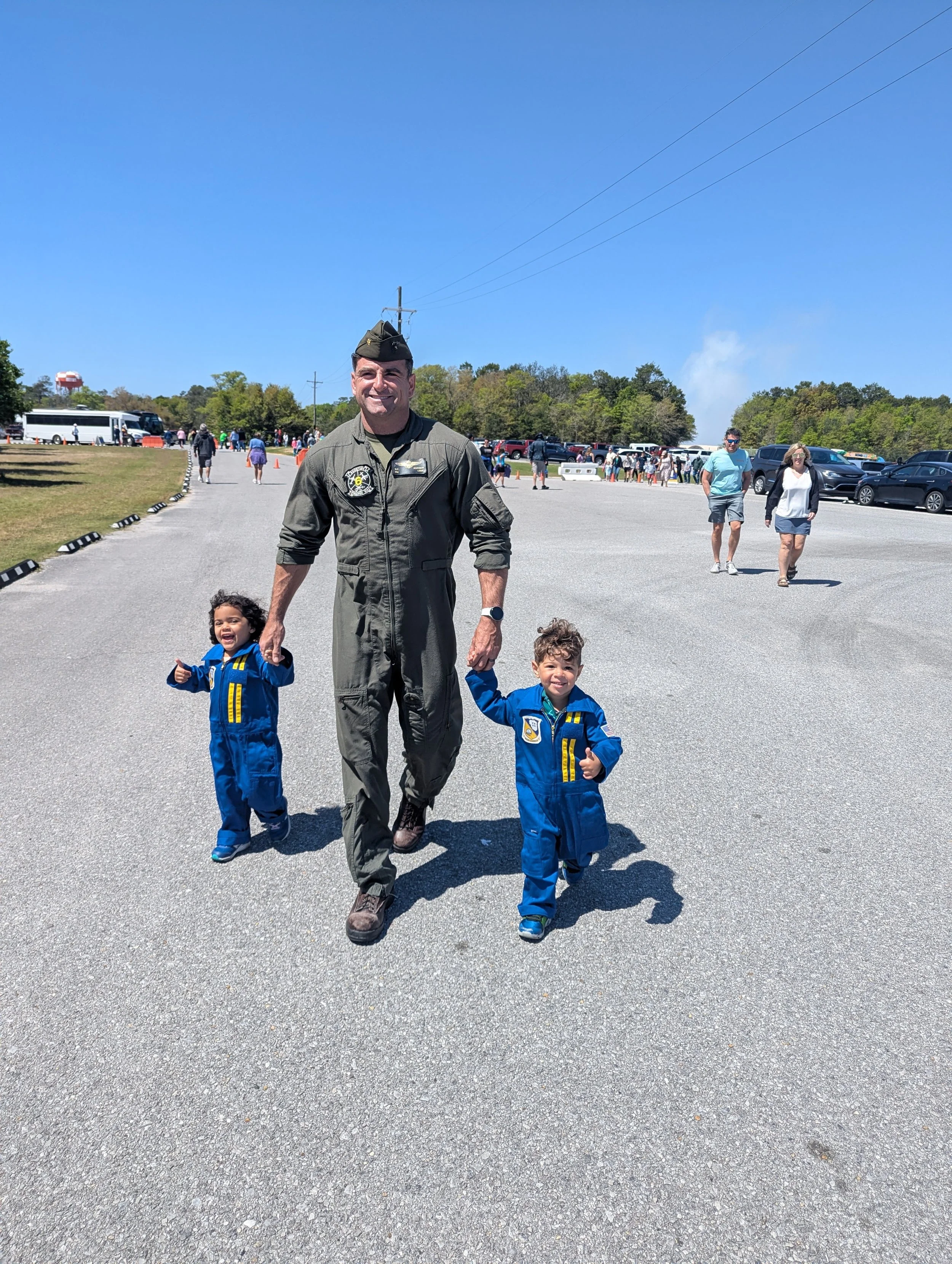A father in military uniform holding hands with two young children, walking on a gravel path at an air show on a sunny day. In the background, there are other people, parked cars, and a line of orange cones.