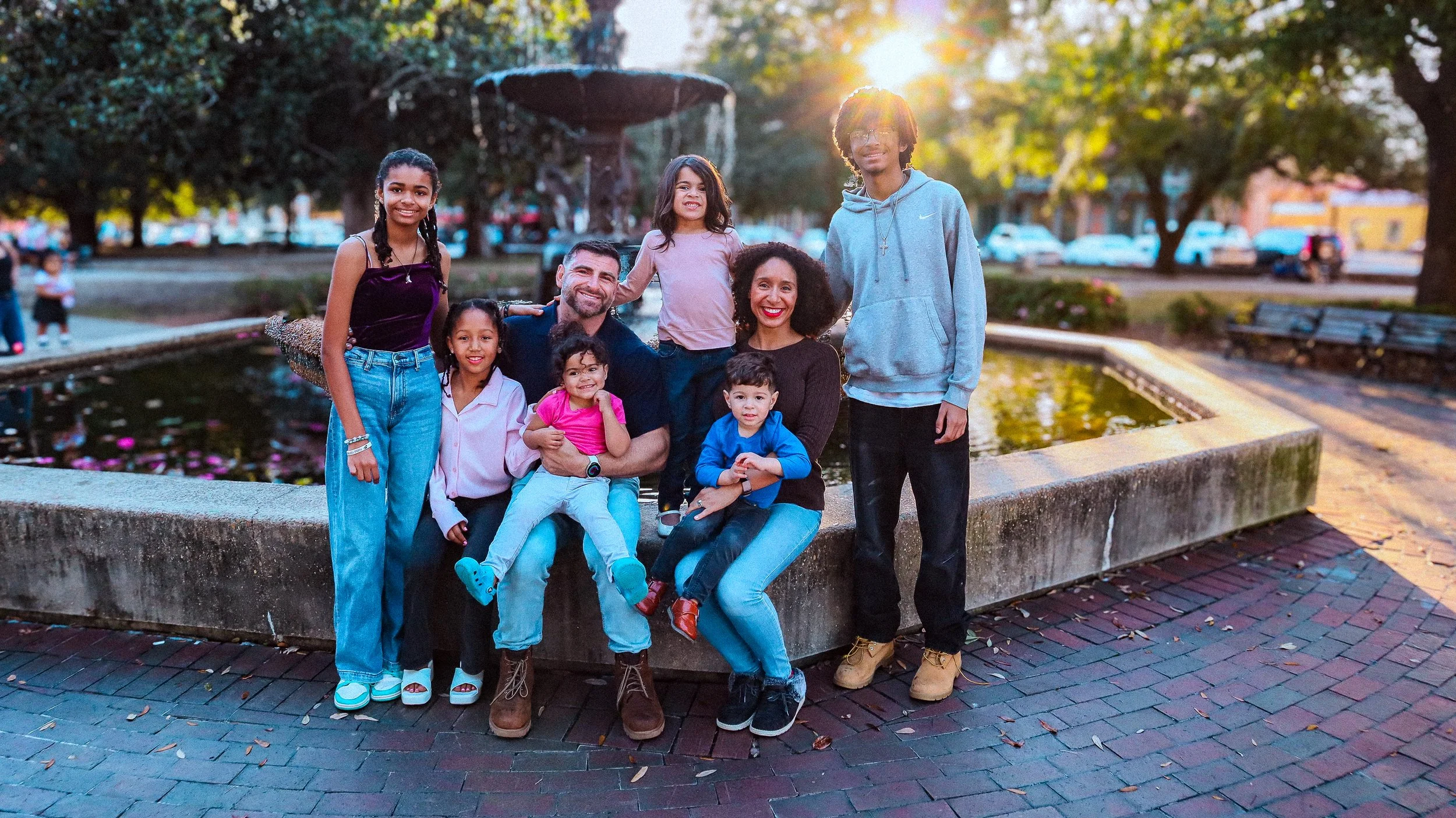 A family of eight people, including children and adults, smiling and posing by a fountain in a park during sunset at Plaza Ferdinand in Pensacola, Florida.