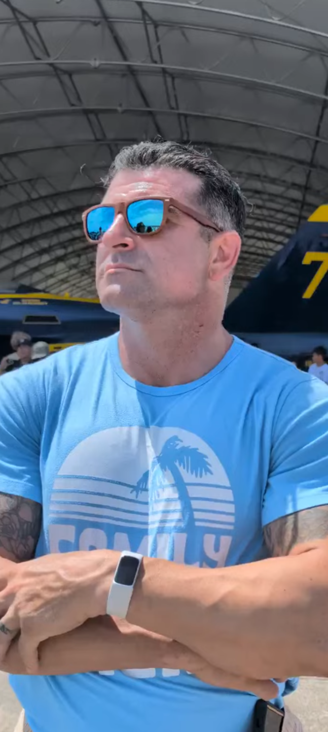 A man with short dark hair, wearing mirrored sunglasses, a light blue t-shirt with a palm tree graphic, and a white smartwatch, standing inside a spacious aircraft hangar with military jets and people in the background watching the Blue Angels fly.
