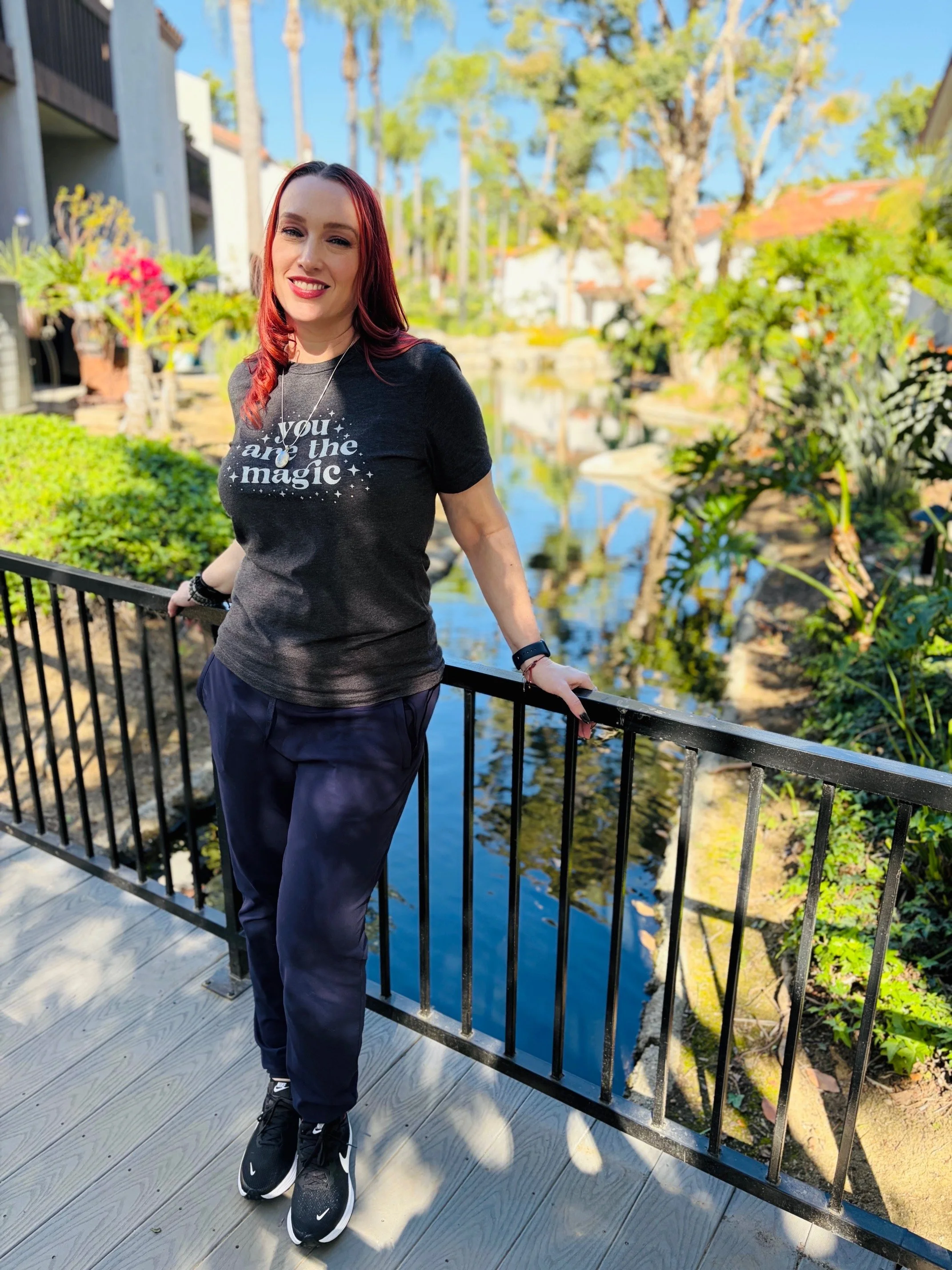 A woman with red hair and a black t-shirt with the words 'you are the magic' stands on a wooden bridge with black railing near a small stream, surrounded by green plants, trees, and houses with blue sky in the background.