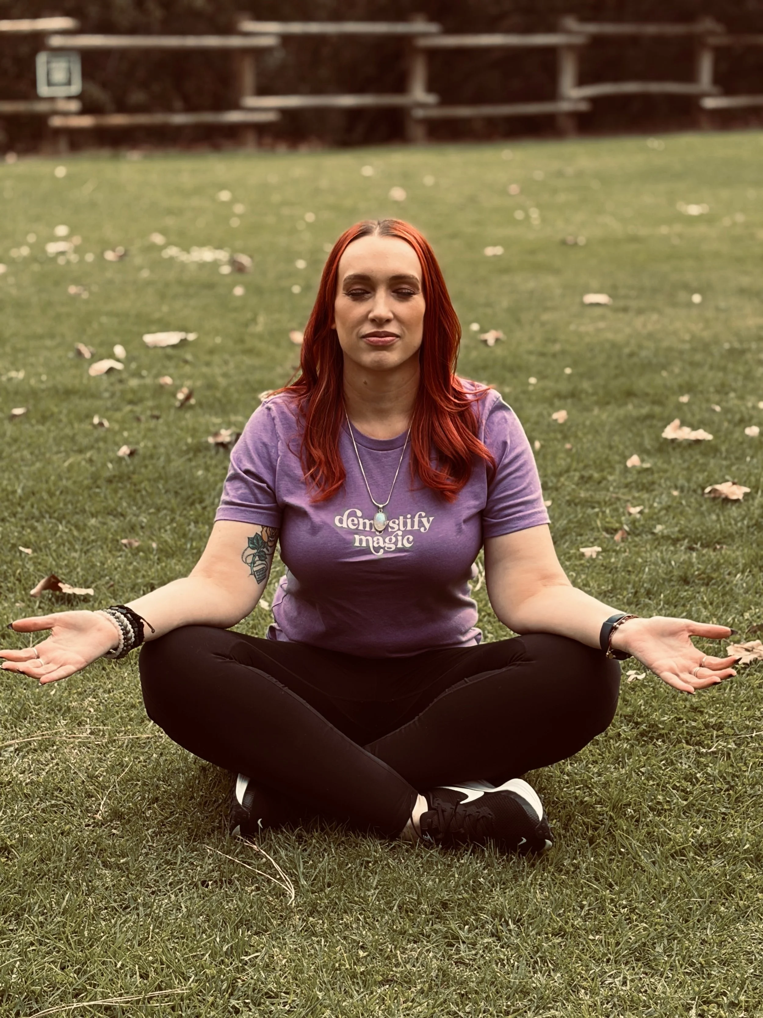 A woman with red hair and tattoos practicing yoga outdoors on a grassy field, seated in a cross-legged meditation pose with eyes closed and hands resting on her knees.