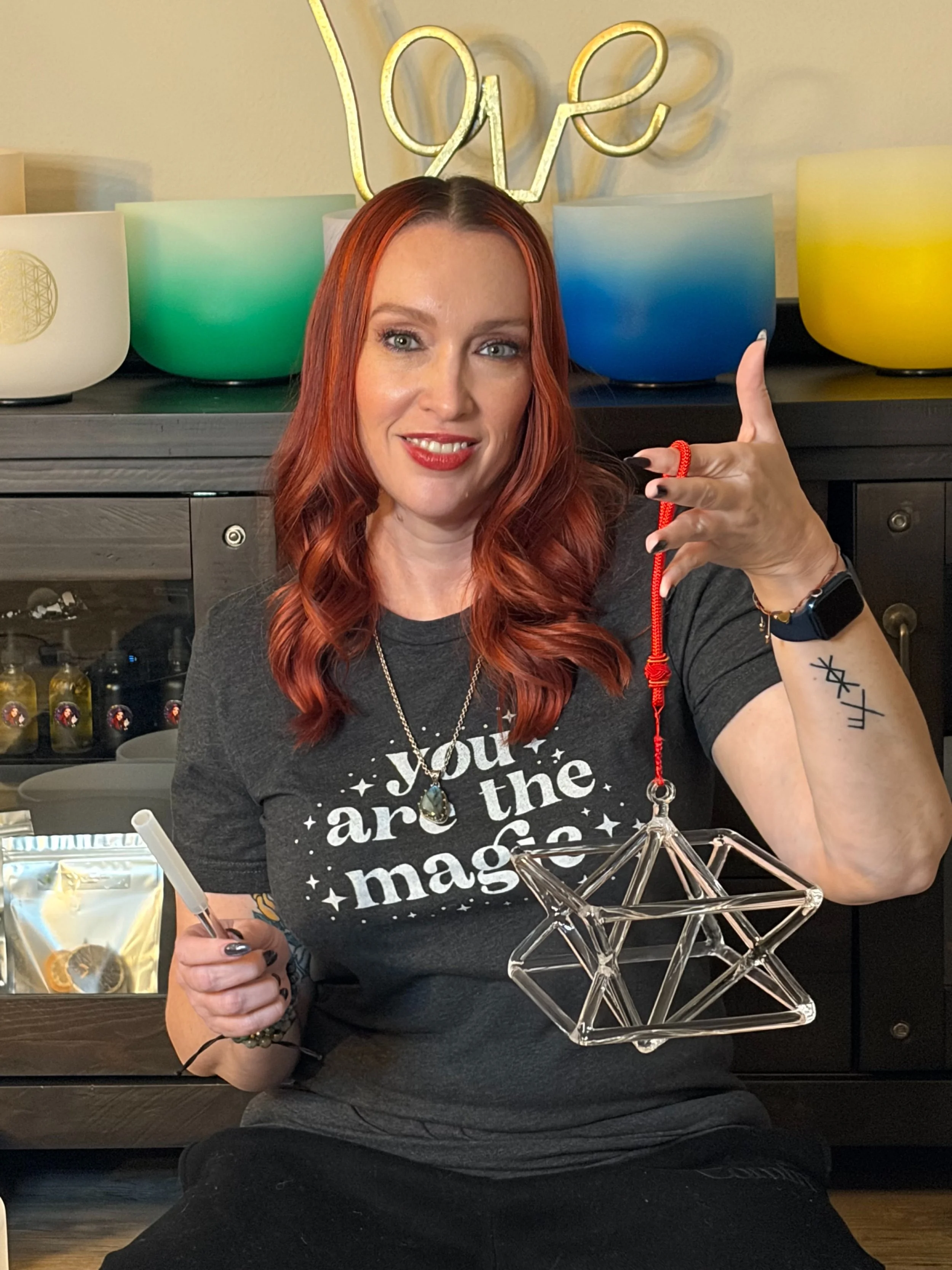 A woman with red hair and tattoos on her arm is sitting at a table, holding a drink and a red string, with a glass star-shaped ornament in front of her. She is smiling with red lipstick and wearing a black t-shirt that says 'You are the magic'. Behind her, there are colorful bowls and a sign that says 'Love' in neon lights.