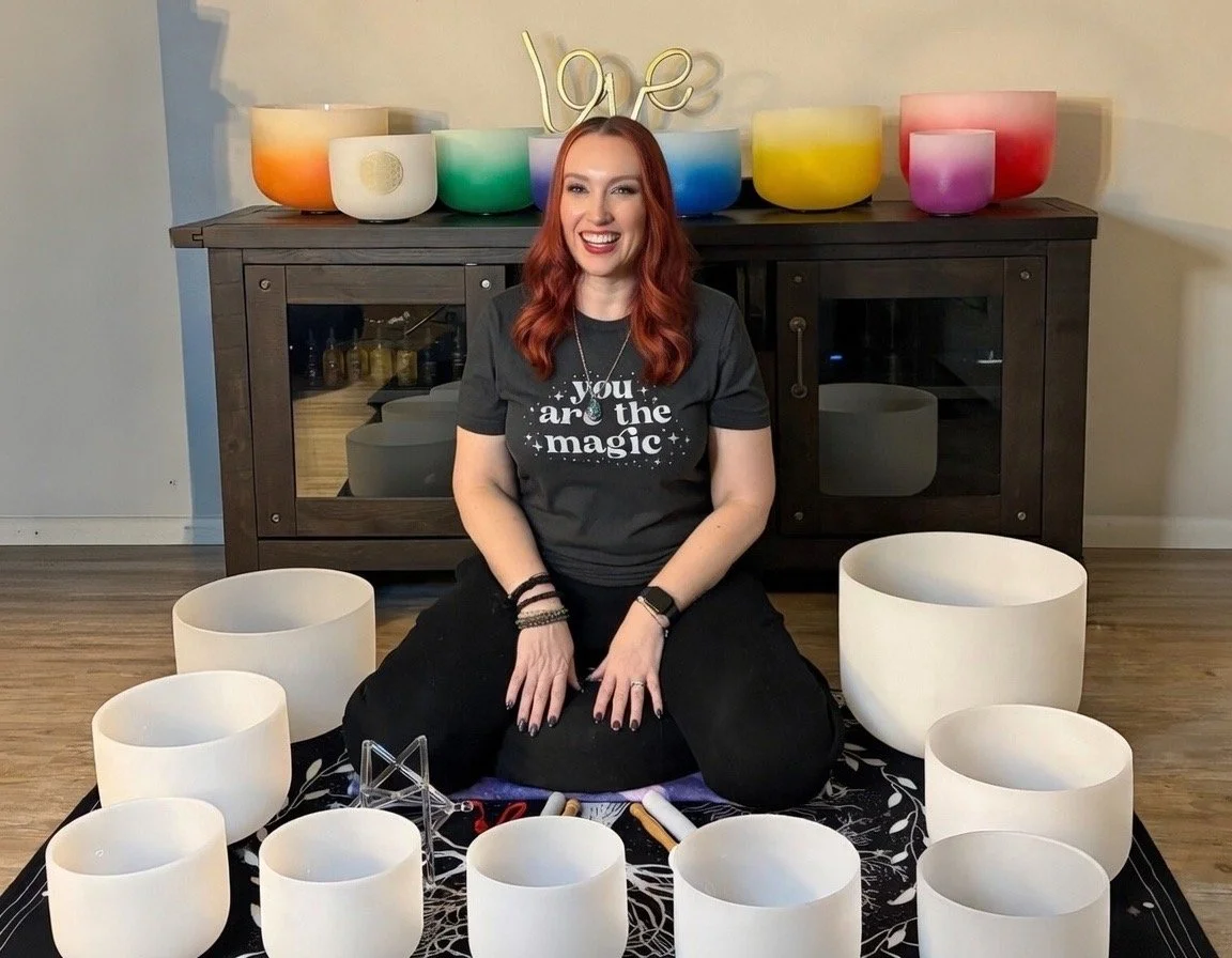 A woman with red hair sitting on the floor in front of a collection of white crystal singing bowls arranged in a semi-circle. She is smiling and wearing a black T-shirt with the message 'you are the magic' and black pants. Behind her is a wooden cabinet with colorful lamps and a decorative gold 'love' sign on top.