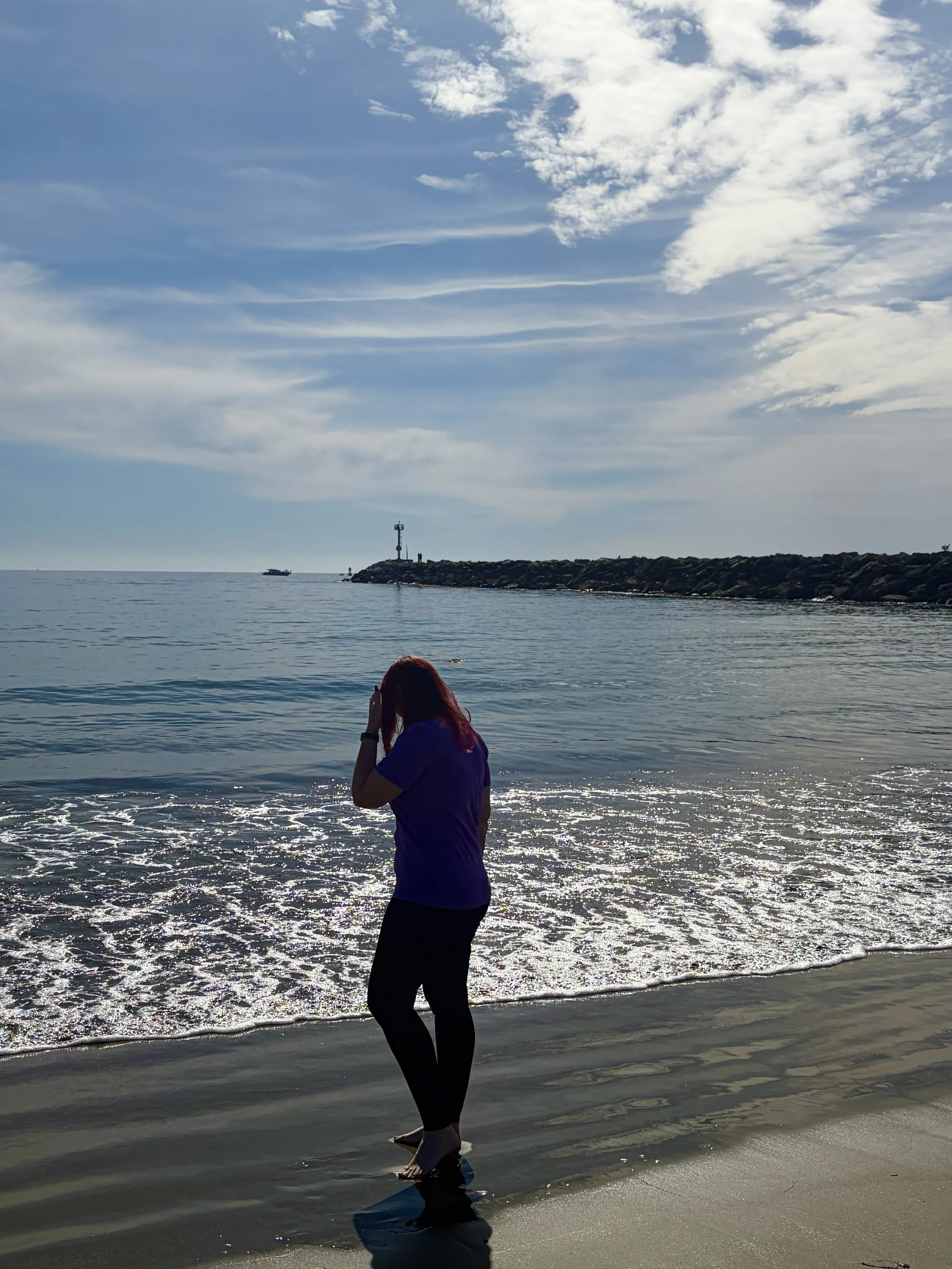 A person with red hair standing at the edge of the water on a beach, facing away, with her hand near her face. The ocean is calm, with a breakwater and lighthouse in the distance under a partly cloudy sky.