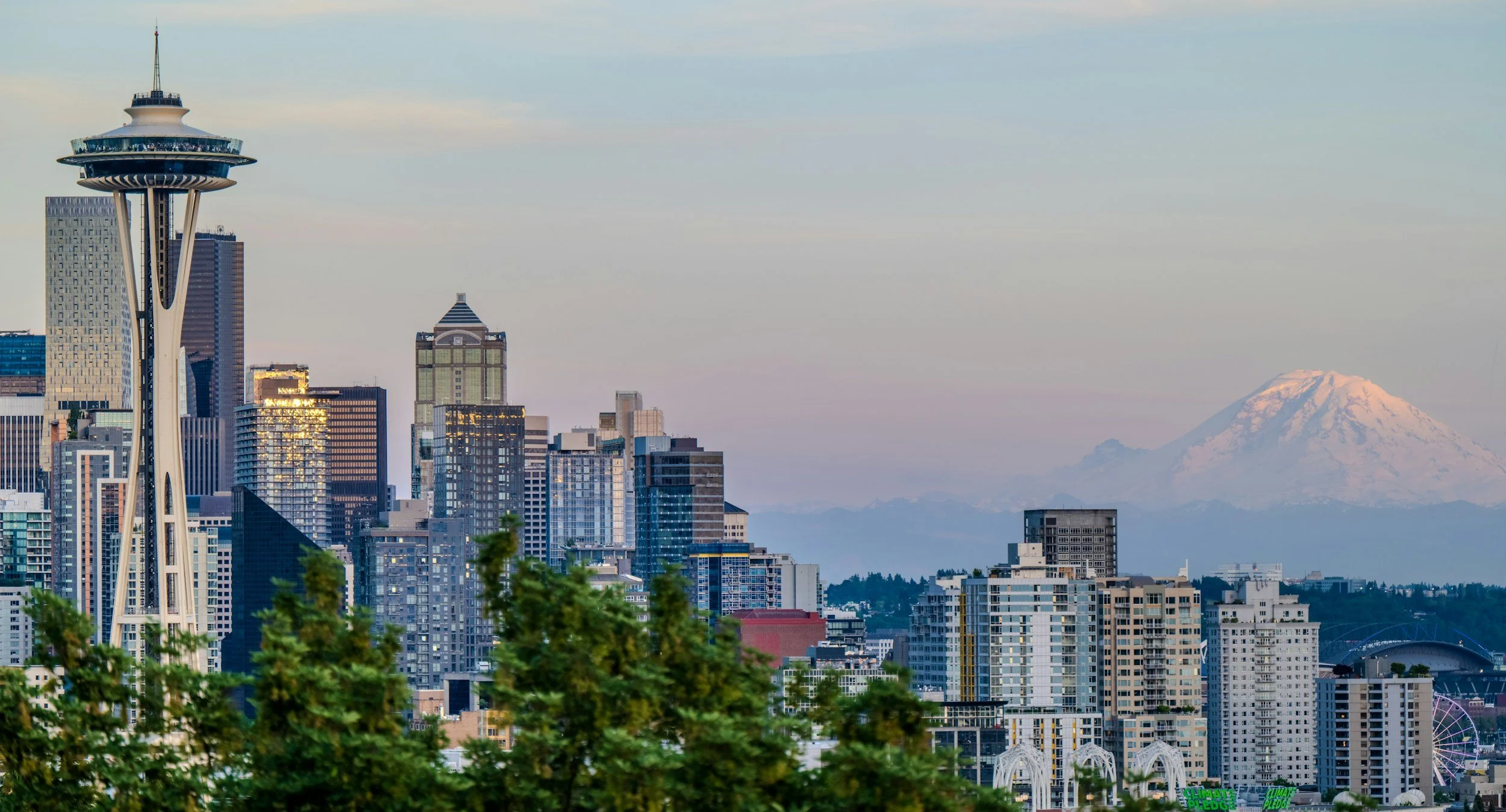 A city skyline featuring numerous tall skyscrapers, the iconic Space Needle, and Mount Rainier in the background on a clear day.